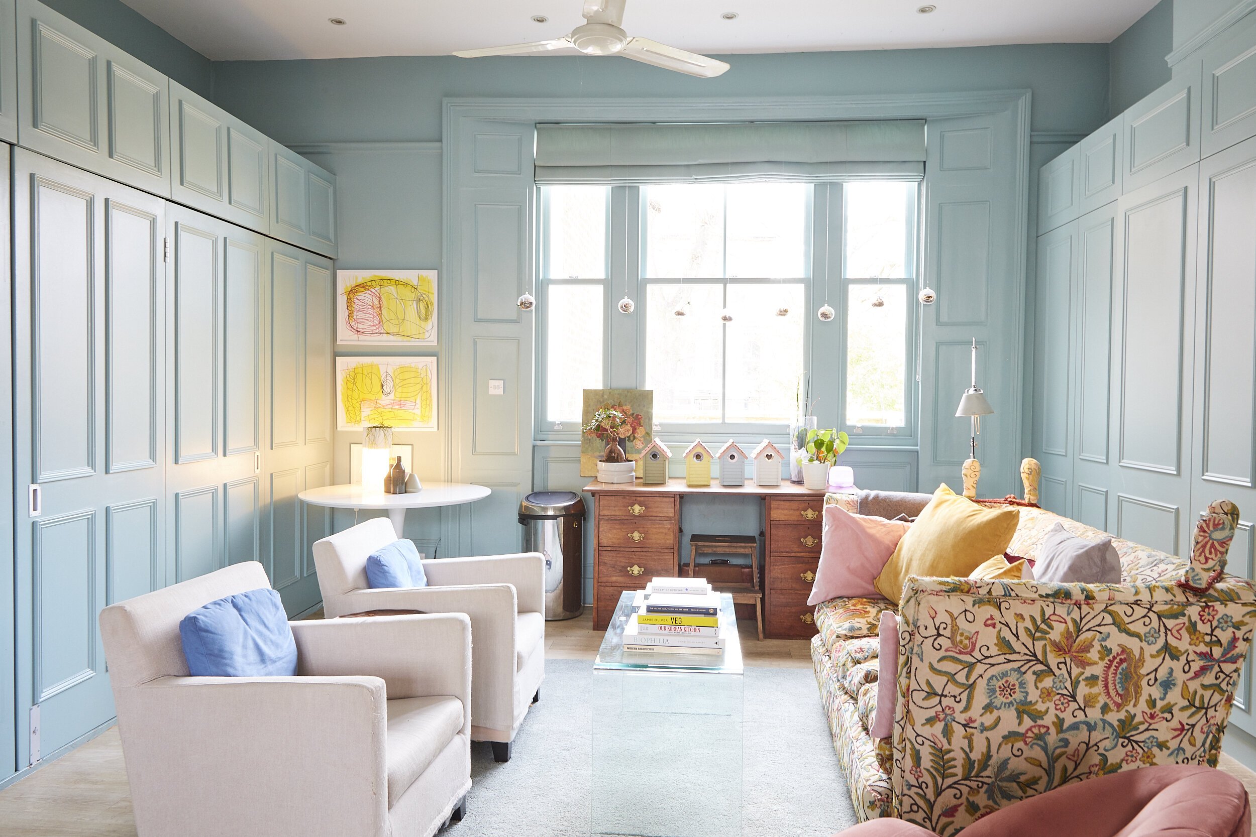 A cozy living room with blue paneled walls, a large window, white armchairs with blue pillows, a floral sofa with colorful cushions, a glass coffee table with books, and various decorations including artwork, birdhouses, and plants.