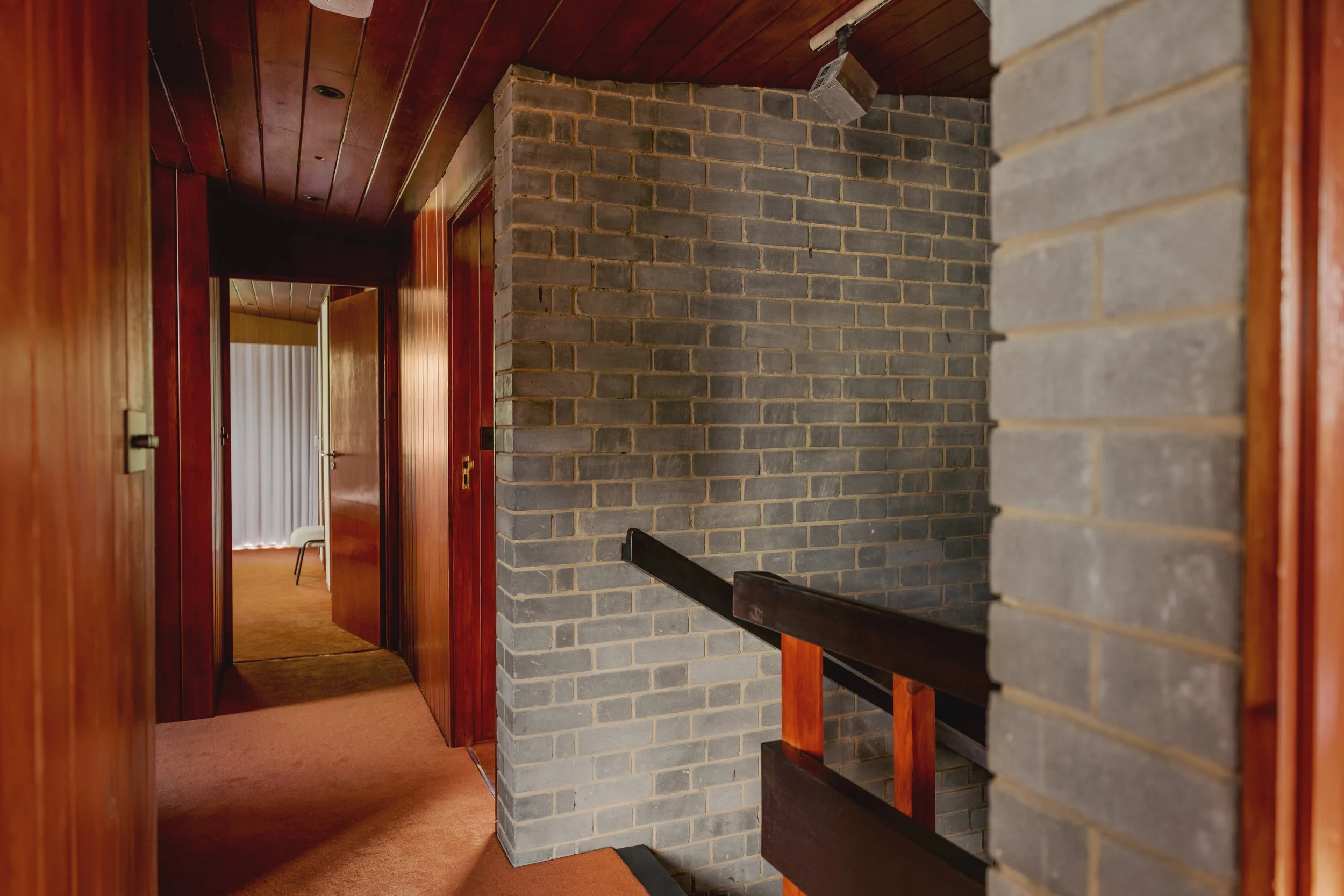 View of an interior hallway with wood-paneled walls and ceiling, a carpeted floor, and a brick wall with a railing on the right, leading to a room with a white chair and vertical blinds.