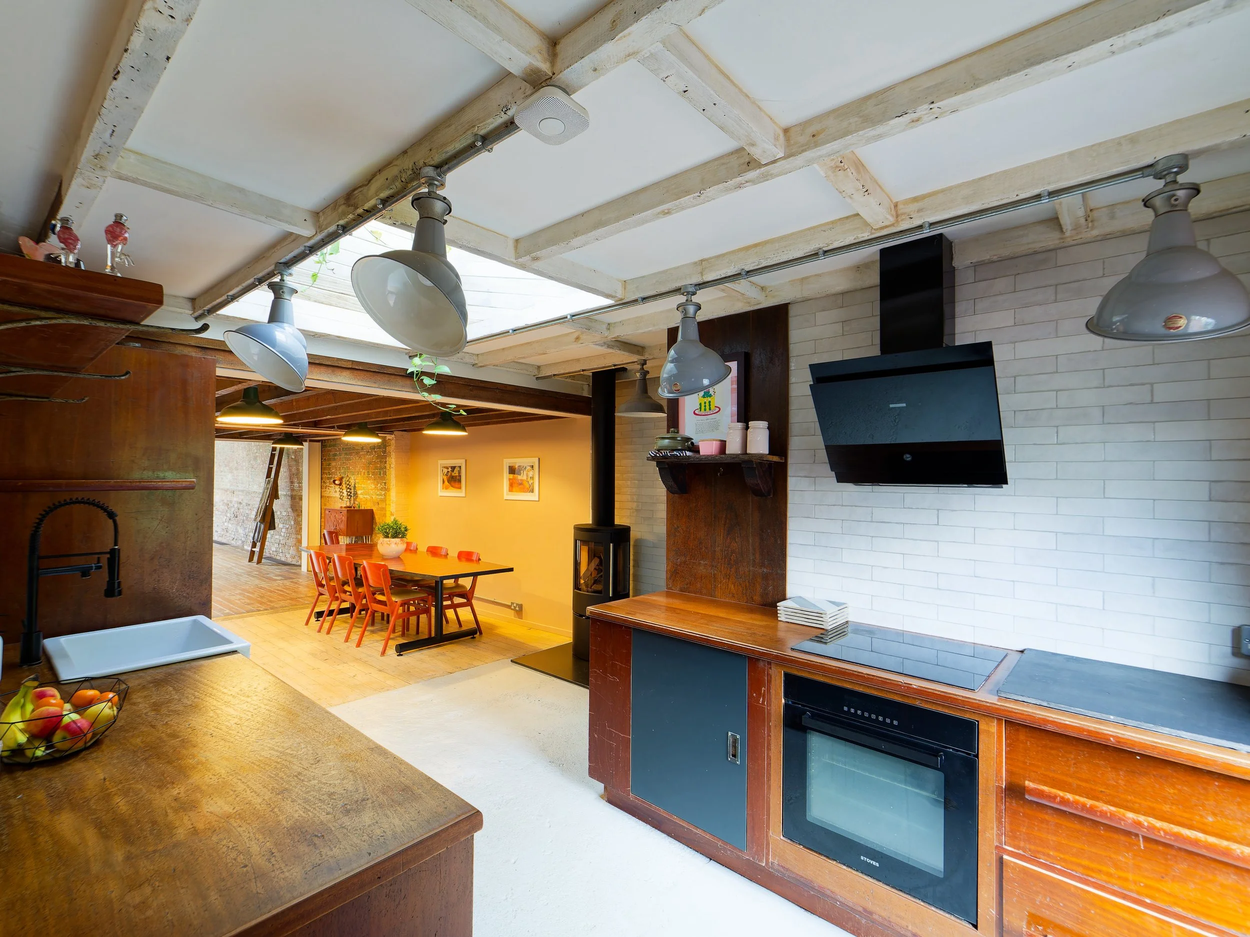 Open kitchen with wooden counters, black oven, white brick wall, ceiling lights, and adjacent dining area with orange chairs and framed artwork