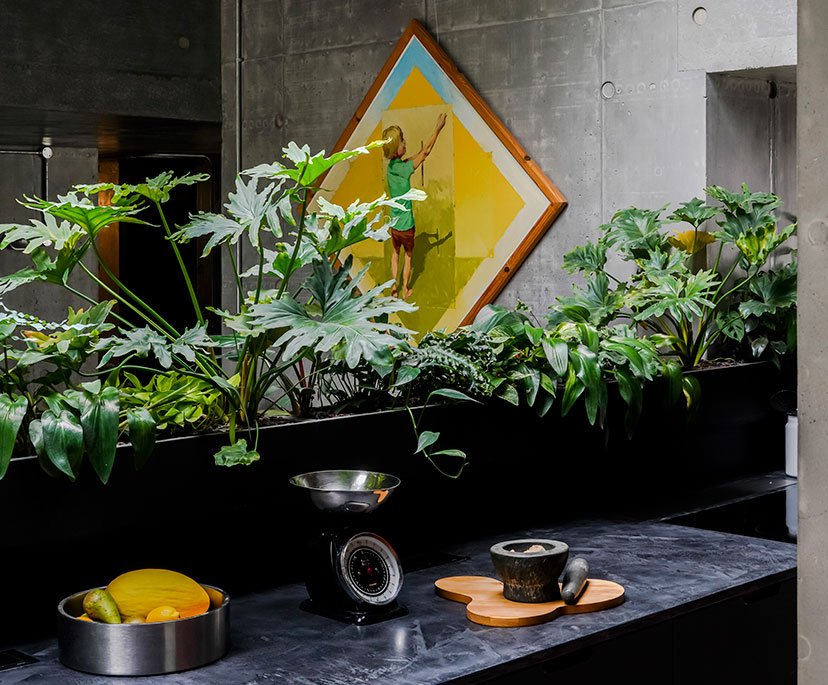 Indoor space with green houseplants in a long black planter, a still life arrangement with a scale, a mortar and pestle, a wooden cutting board, and a bowl of fruit, and a colorful painting of a child climbing a yellow wall in the background.
