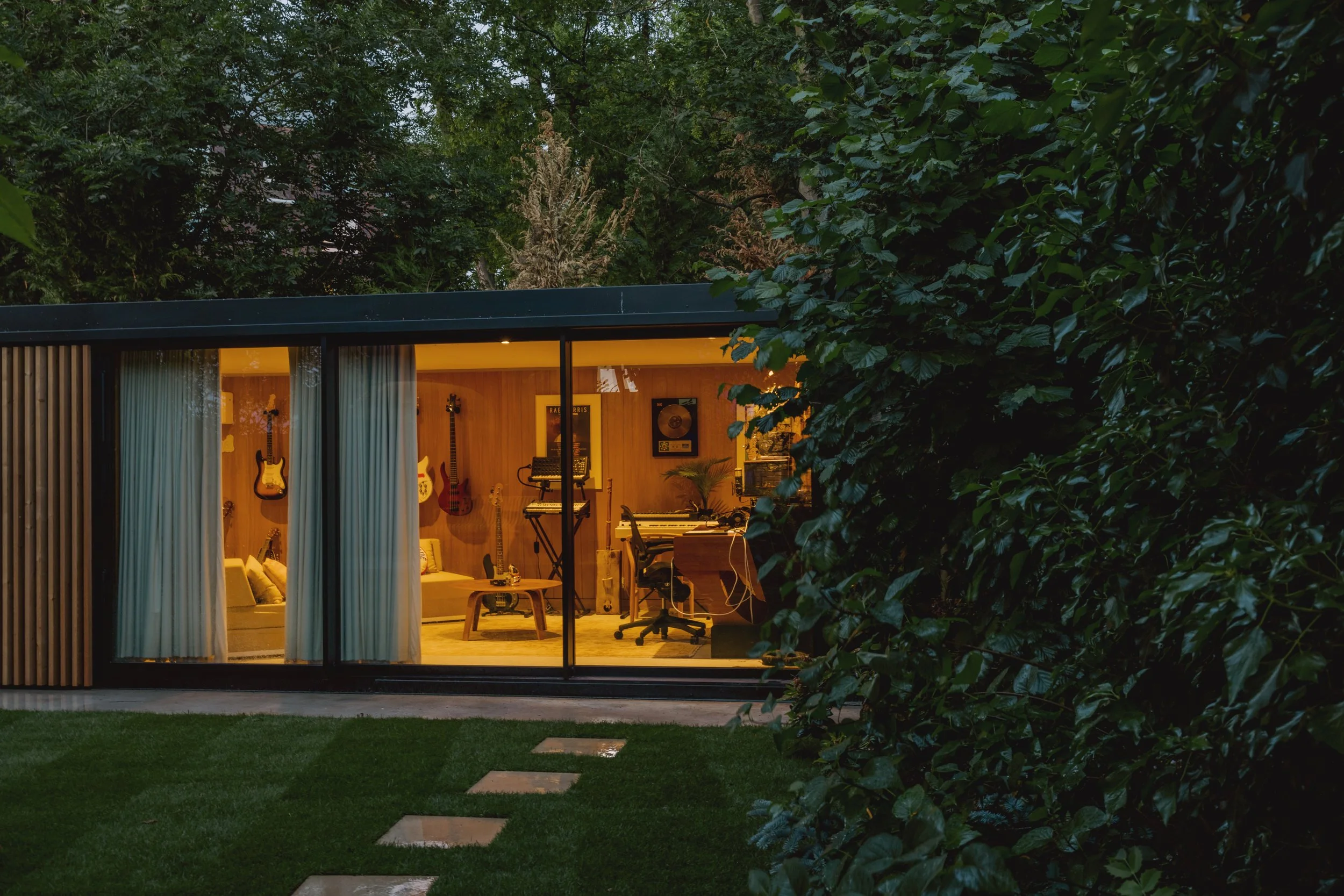 View of a modern house with large glass windows revealing a music studio inside, with guitars on the wall, a keyboard, and a reclining chair, surrounded by lush greenery.