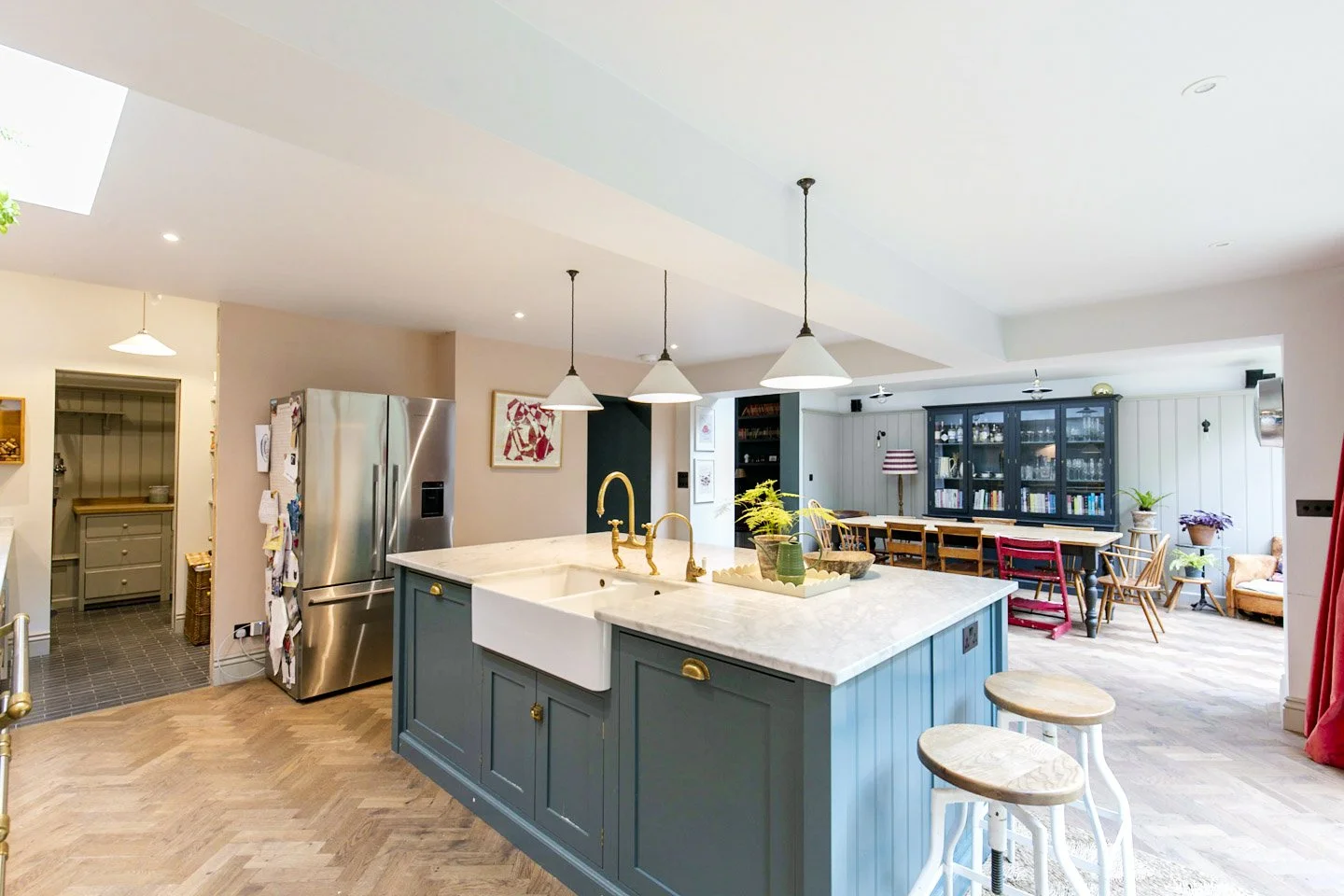Open-concept kitchen with a blue island, marble countertop, gold fixtures, and a dining area with chairs and a black bookcase in the background.