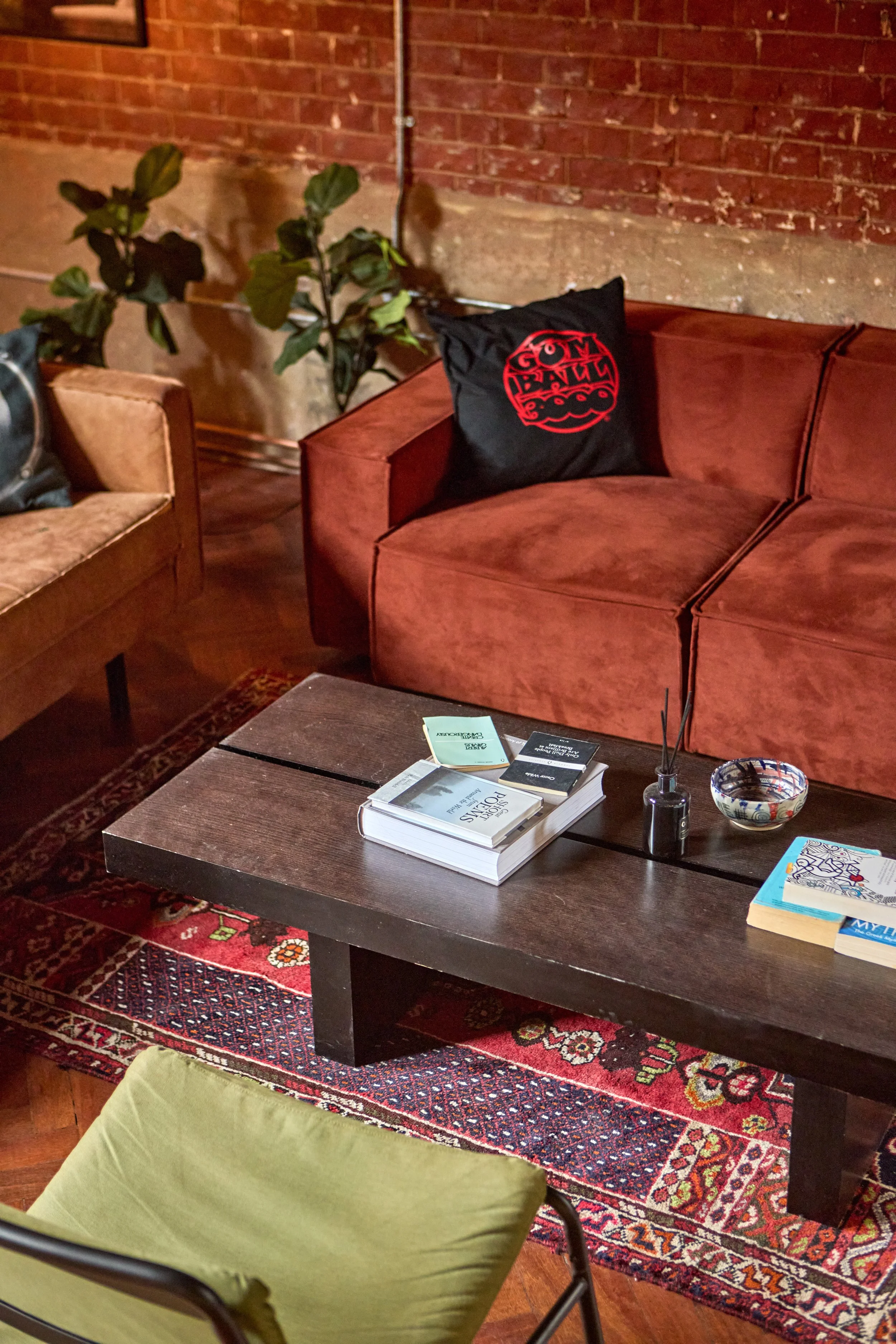 A cozy living room with a reddish-brown velvet sofa, a black pillow with a red design, behind a dark wooden coffee table with books, a small reed diffuser, and a decorative bowl on a patterned rug. There is a brick wall and some green plants in the b