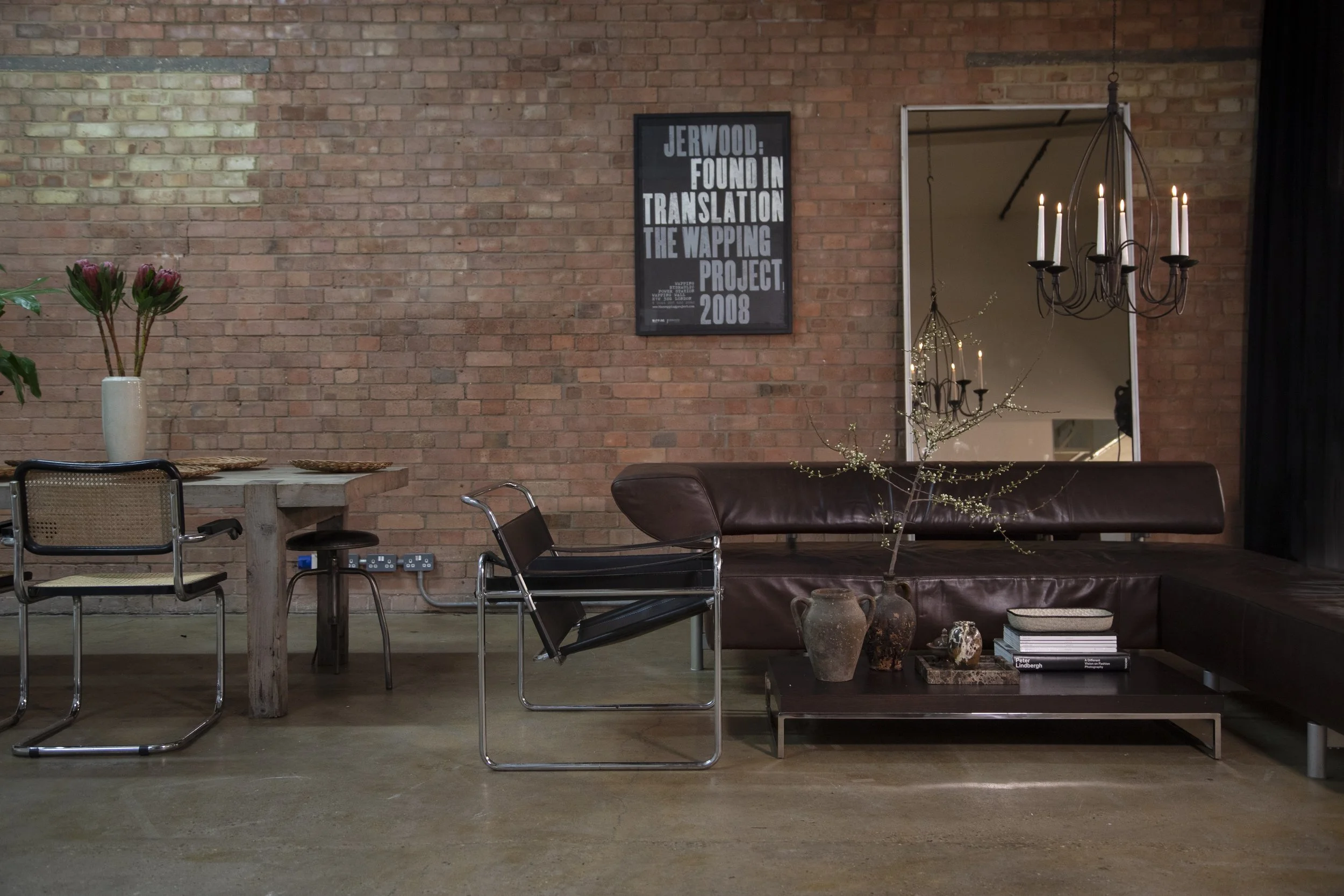 Living room with brick wall, black-framed poster, mirror, chandelier, leather sofa, flowers, and modern chairs.