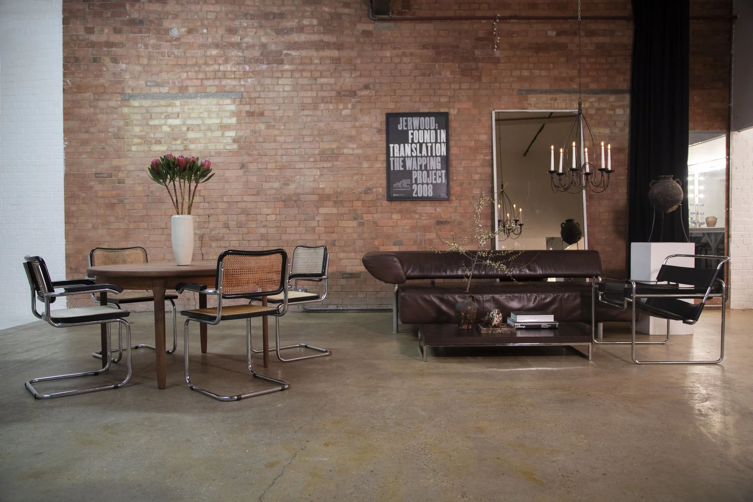 Modern interior with a brick wall, a round wooden table with six chairs, a tall vase with pink flowers, a leather couch with decorative vases and books, a mirror, and chandeliers with lit candles.