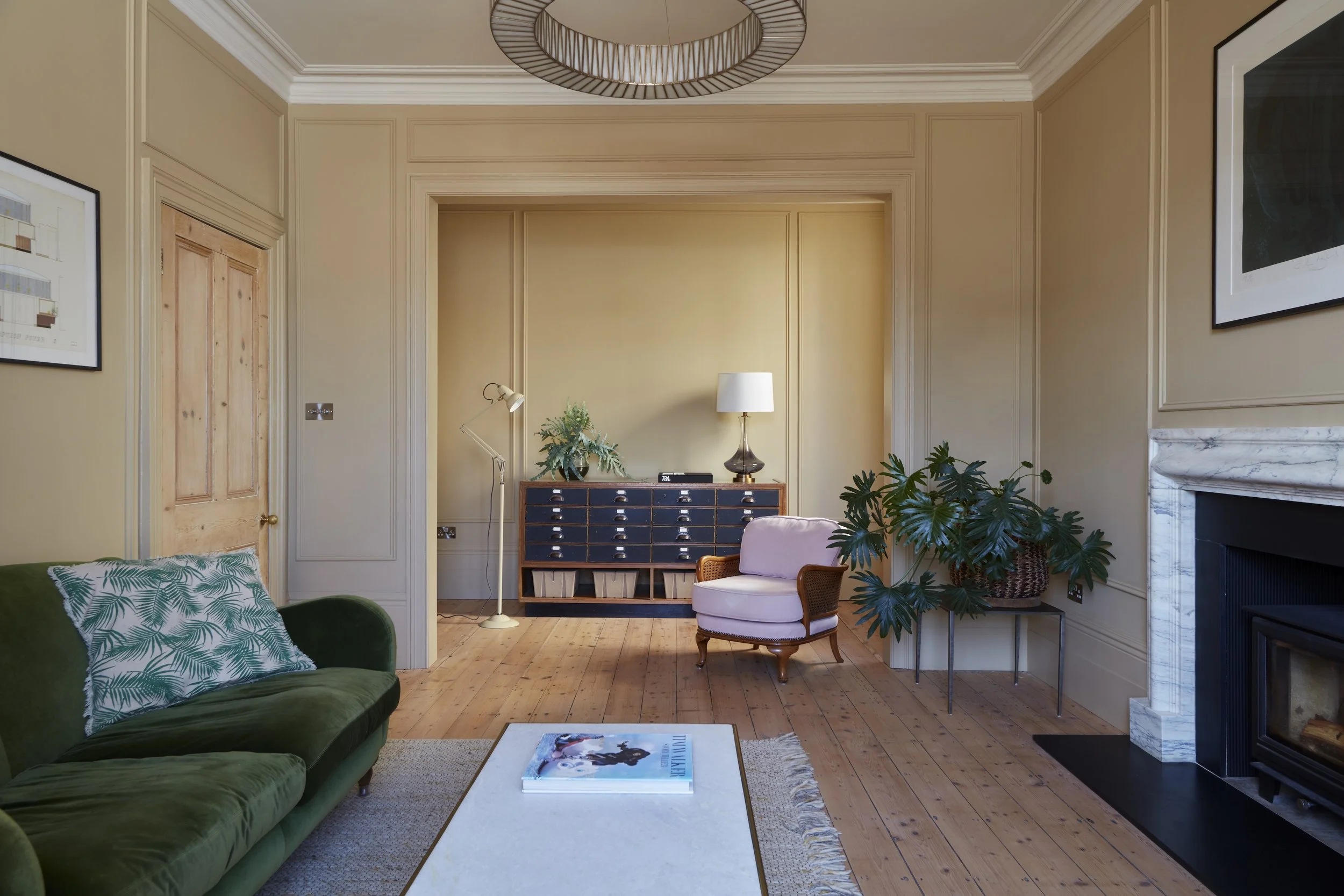 A cozy living room with beige walls, hardwood floors, a green velvet sofa with patterned pillows, a white rug, a low white coffee table with a magazine, a small pink armchair, a wooden side table with a plant, a black sideboard with a lamp, a floor l