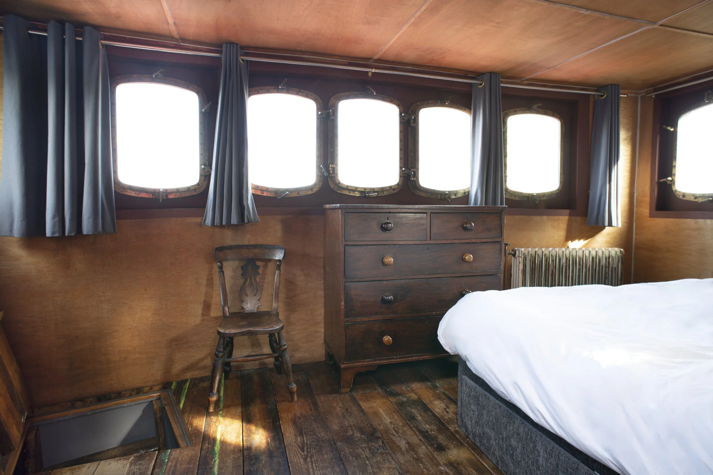 Interior of a rustic bedroom with wooden walls, five windows with blue curtains, a wooden chair, a dark wooden dresser, and part of a bed with white sheets.