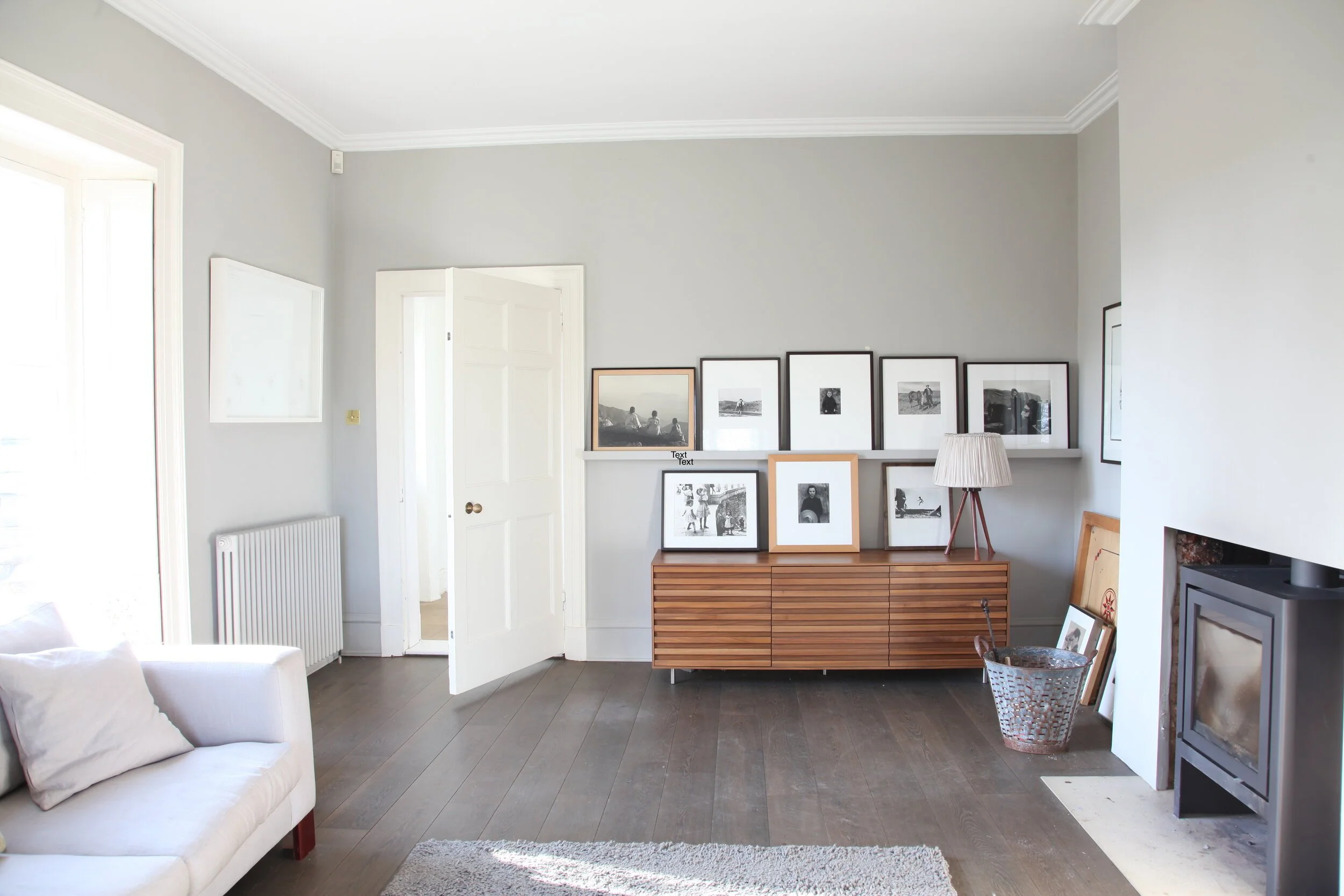 Living room with light gray walls, dark wood floor, white door open to another room, radiator, white sofa with pillow, wooden entertainment unit with framed photos, small table with lampshade, basket, and additional framed photos near fireplace.