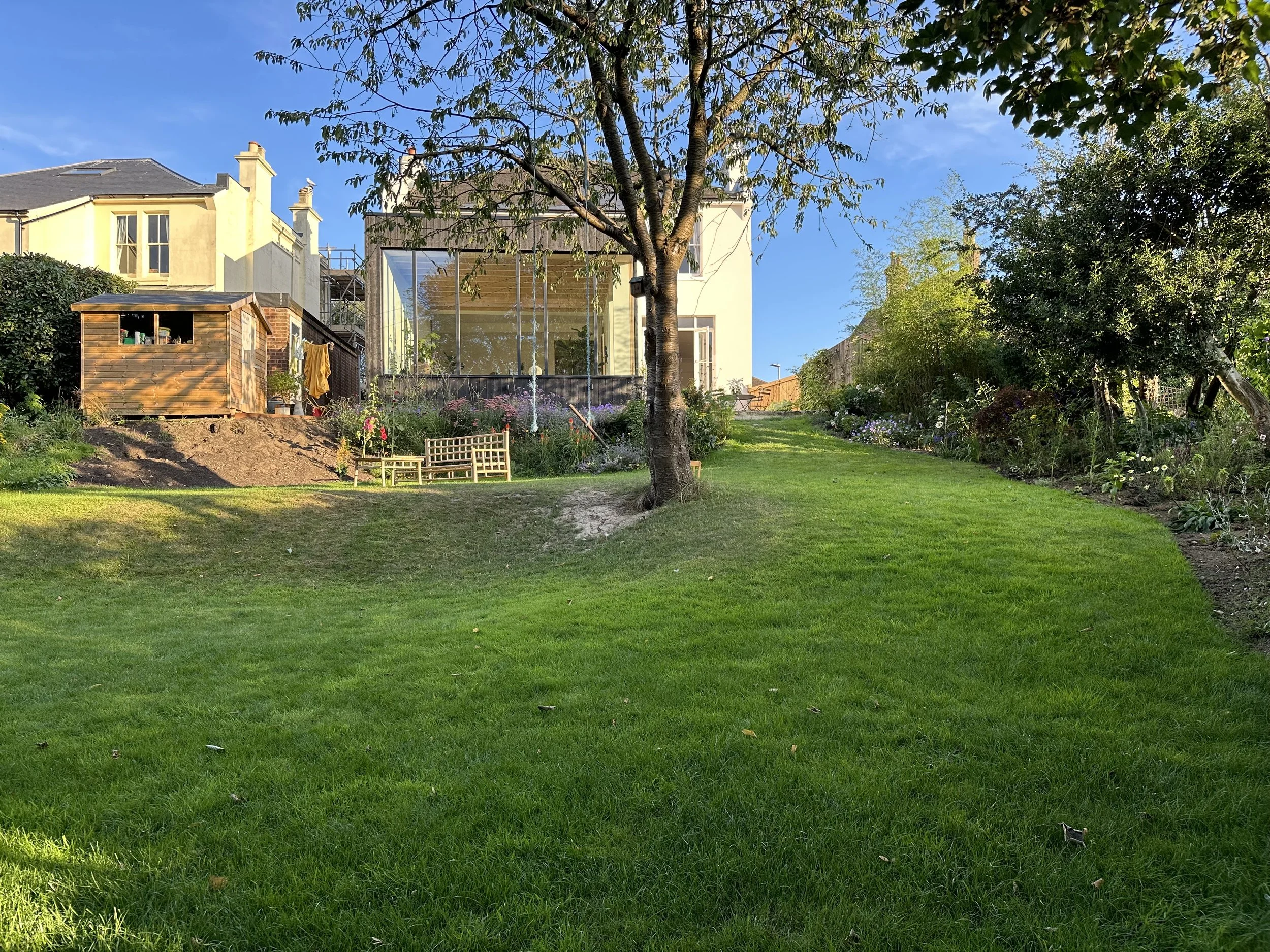 A backyard with green grass, a tree in the center, a garden area on the right, a wooden shed on the left, and a modern glass house in the background under a blue sky.