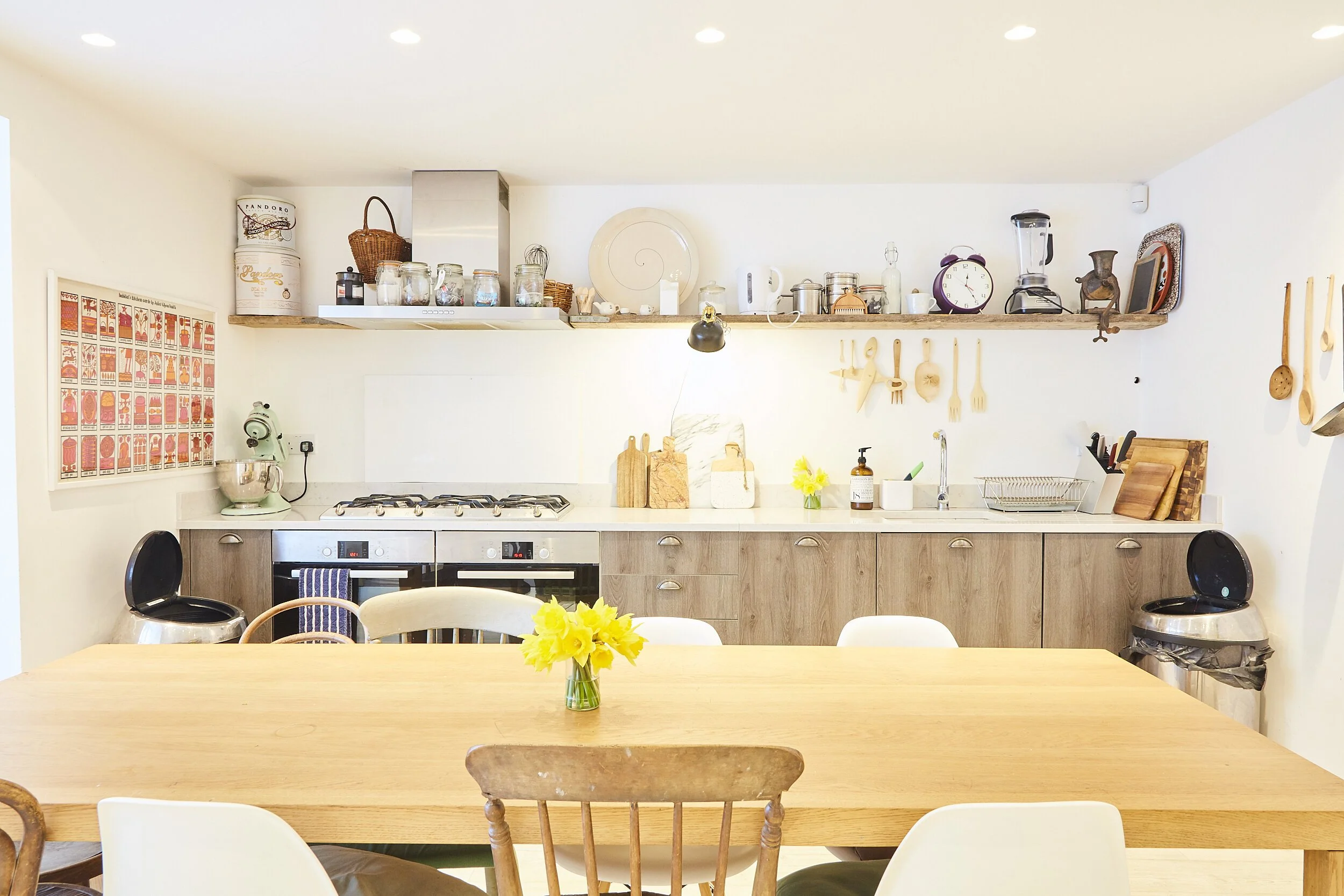 Bright kitchen with wooden table and chairs, open shelves with kitchenware, yellow flowers in a vase on the table.