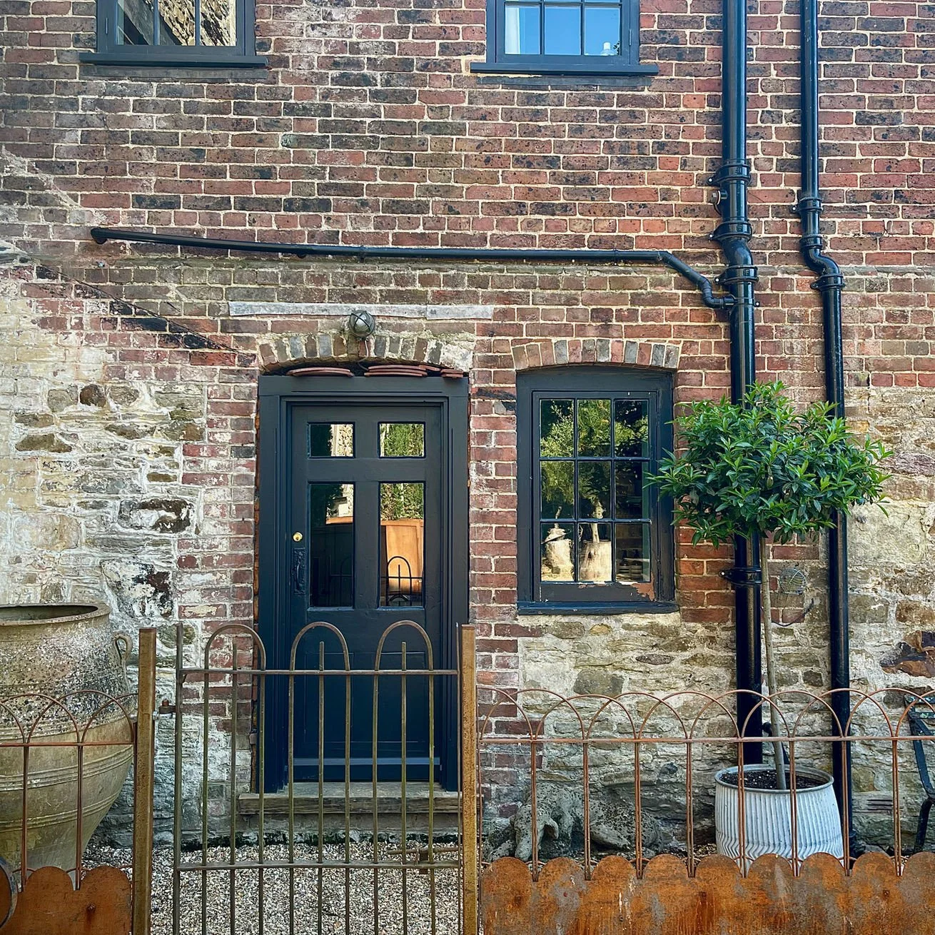 A brick building exterior with a black door and a window, black drainpipes, a small tree in a white pot, and a metal fence in front.