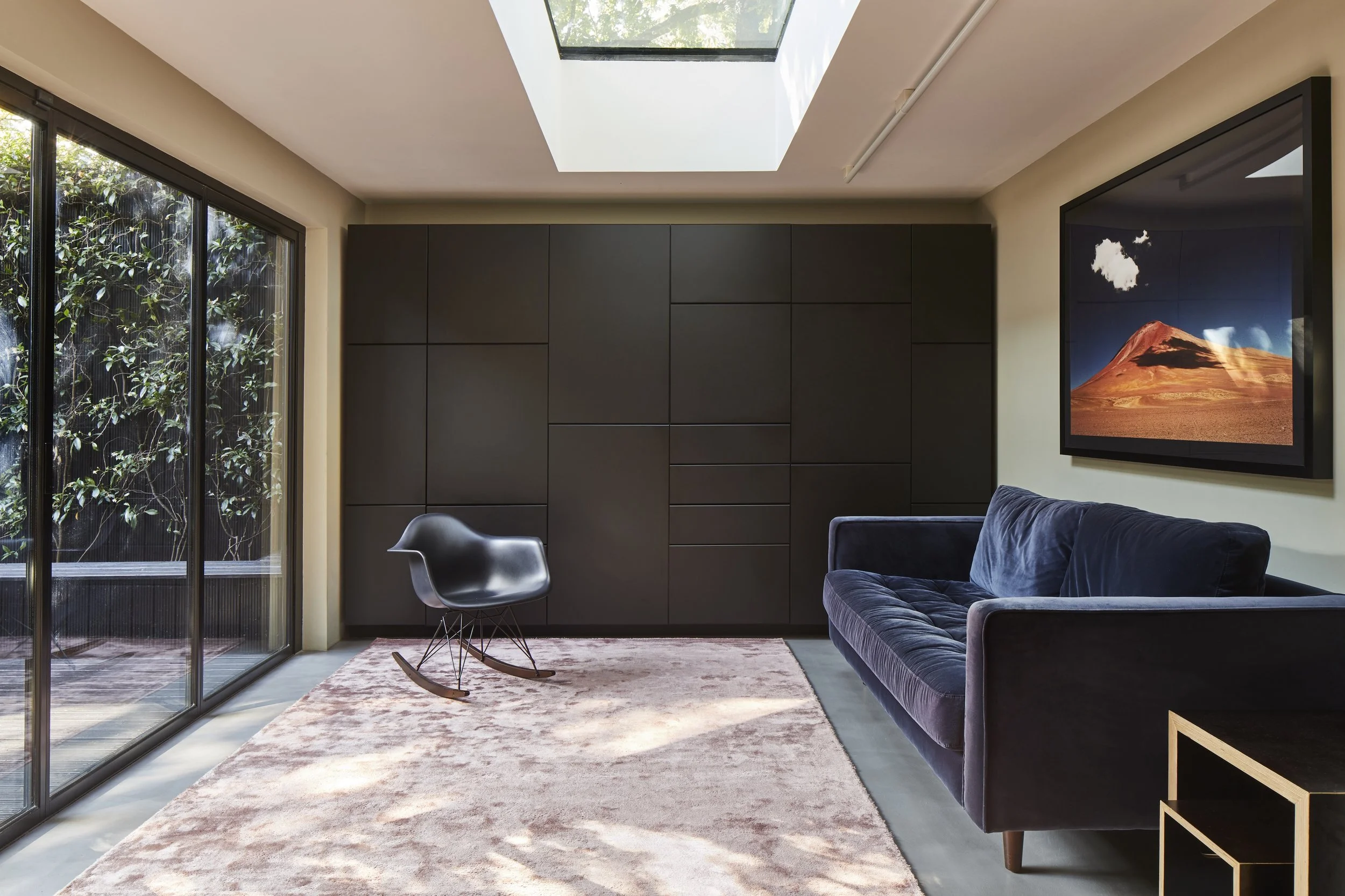Modern living room with large sliding glass doors, black cabinet wall, framed landscape photograph, blue velvet sofa, black rocking chair, pink area rug, and skylight.
