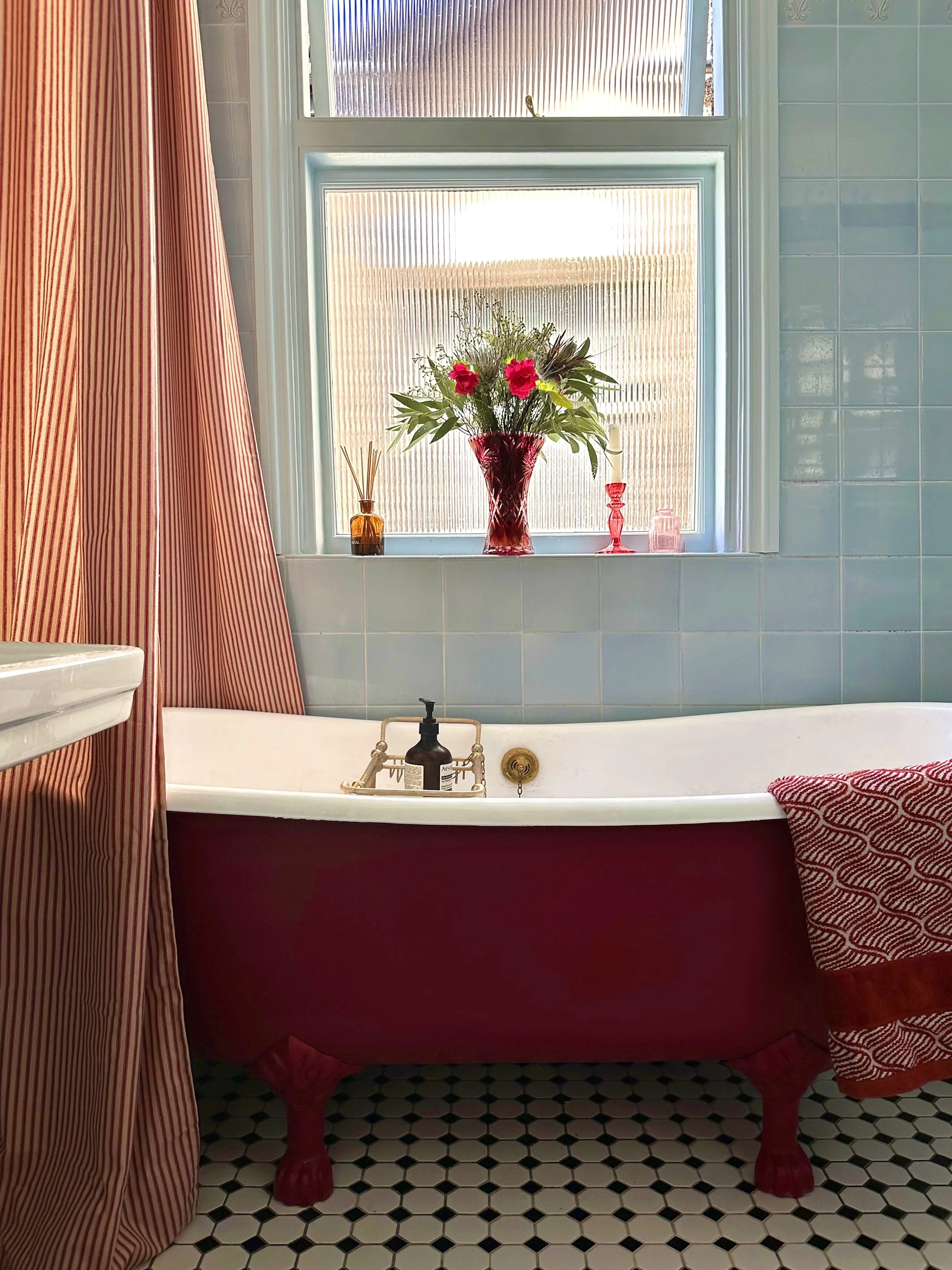 A vintage red clawfoot bathtub in a bathroom with a window. The window has glass blocks and is decorated with a large red vase of flowers and small pink decorative objects. The bathtub has a soap dispenser and a red and white striped towel on the edg