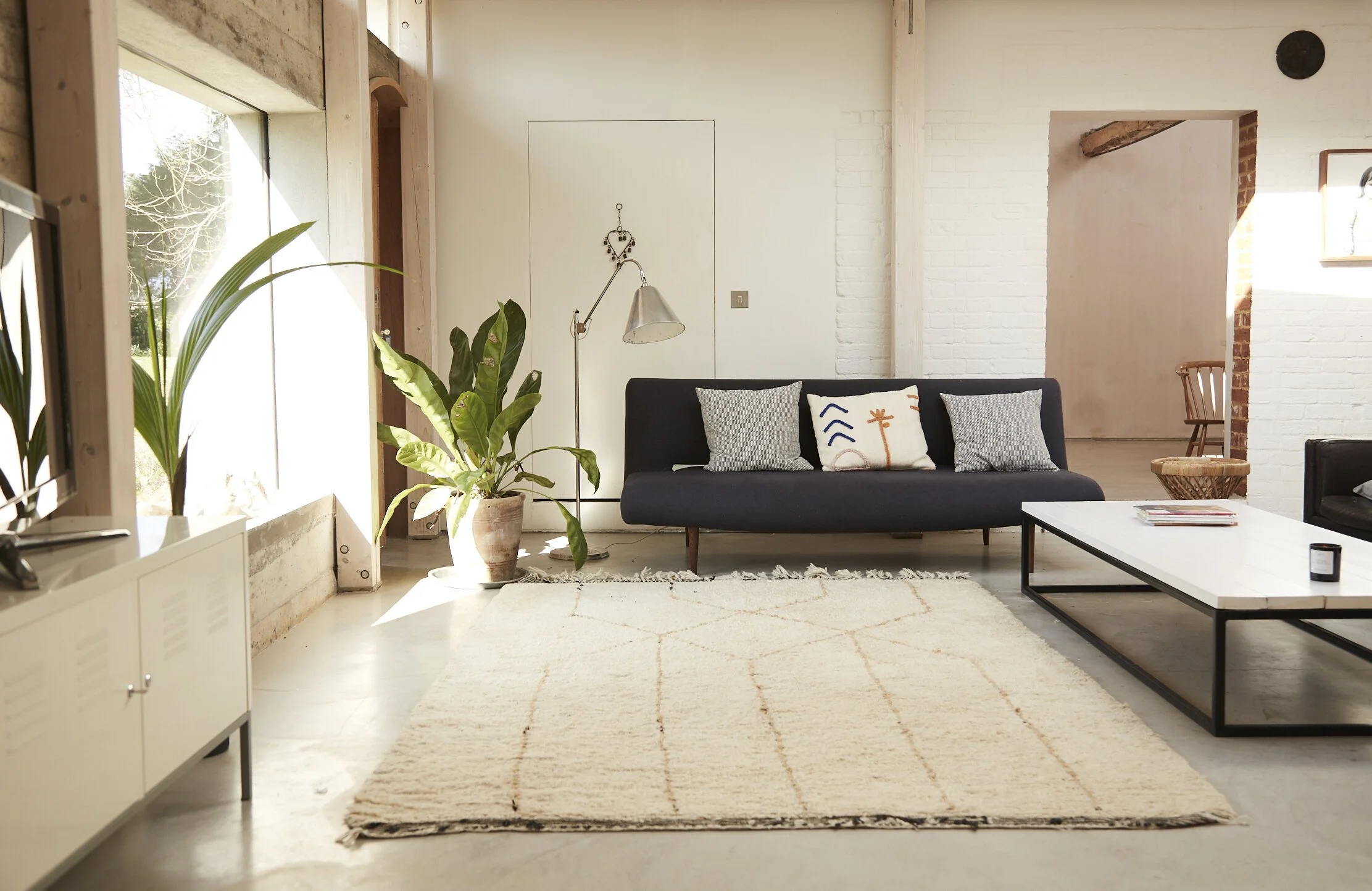 Living room with large window, black sofa, white cushions, a white rug with a diamond pattern, a potted plant, and a white coffee table.
