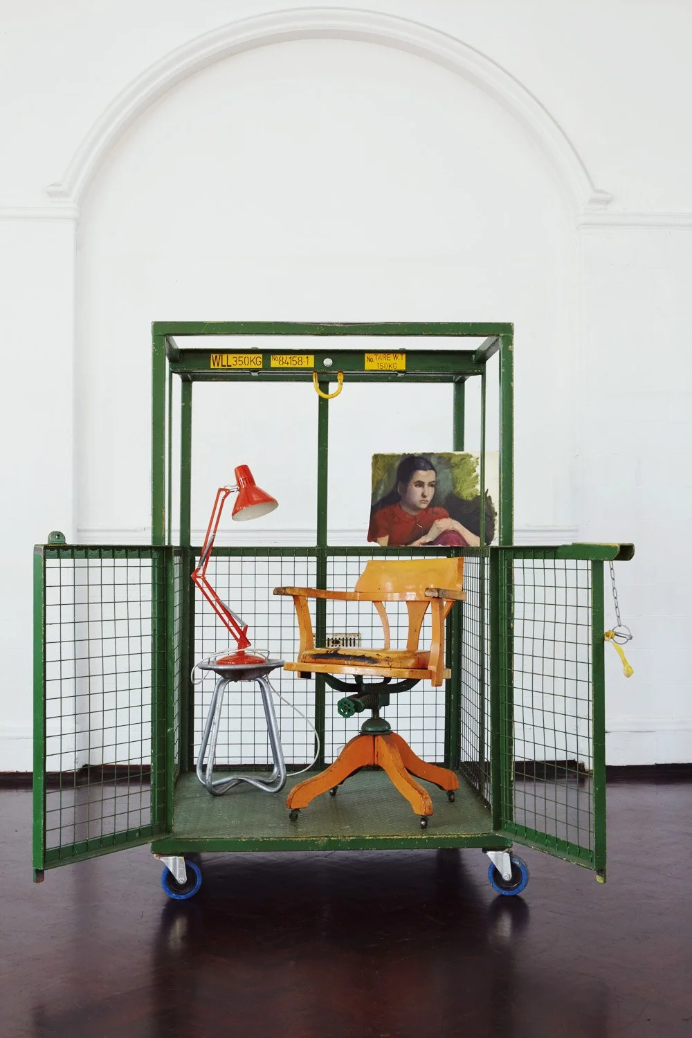 An art installation featuring a green metal cart with a cage-like enclosure, containing an orange office chair, a red desk lamp, a small stool, and a portrait of a woman in a red top. The background is a plain white wall with an arched alcove.