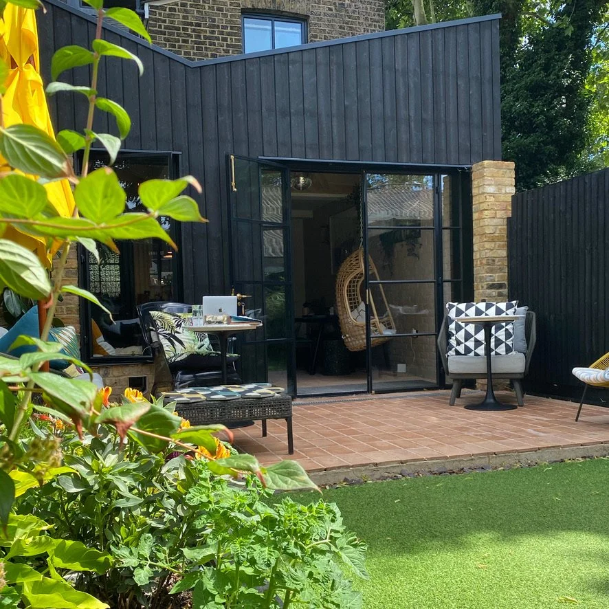 Backyard patio area with seating, a table, and plants, enclosed by black fence with a modern black shed and a brick wall.