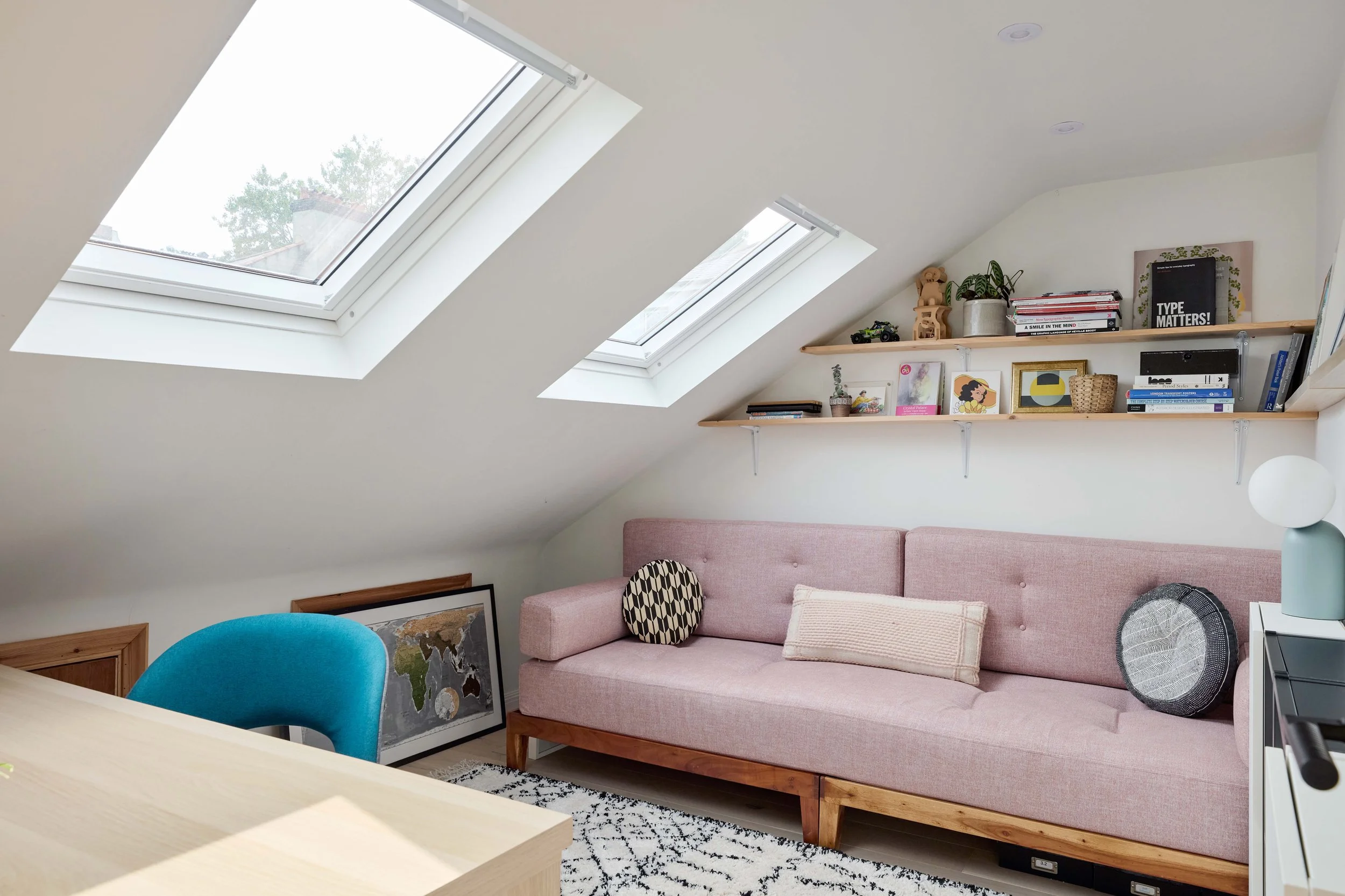 Living room with two large skylight windows, a pink sofa with a striped pillow, a black and white patterned cushion, a black circular cushion, two wooden floating shelves with books and decorative items, and a colorful rug on the floor.
