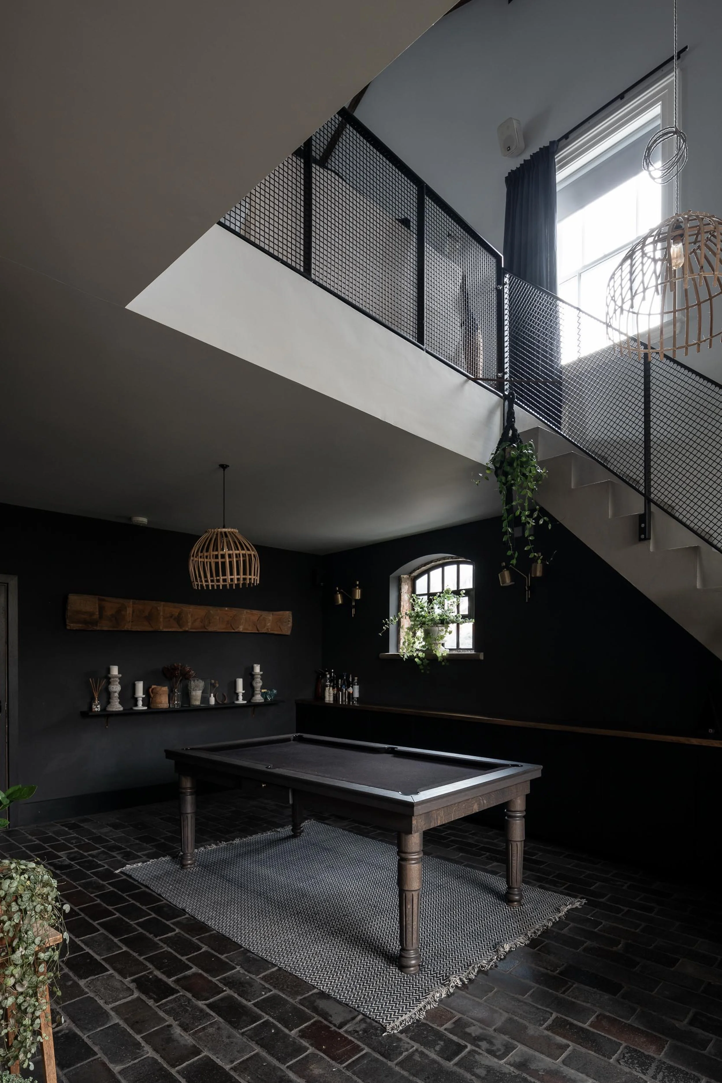 Room with dark walls, pool table on a woven rug, small window with plants, black brick flooring, hanging wicker lights, and balcony with black metal railing.