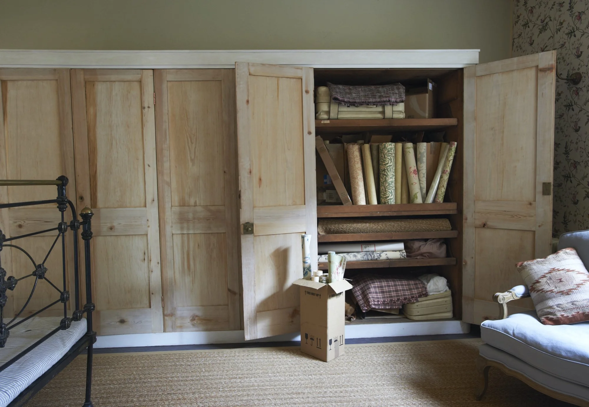 A partially open large wooden closet filled with rolls of wallpaper and fabric, with a cardboard box in front containing wallpaper rolls, in a room with beige carpet, a black metal bed frame, and a white armchair with a patterned cushion.