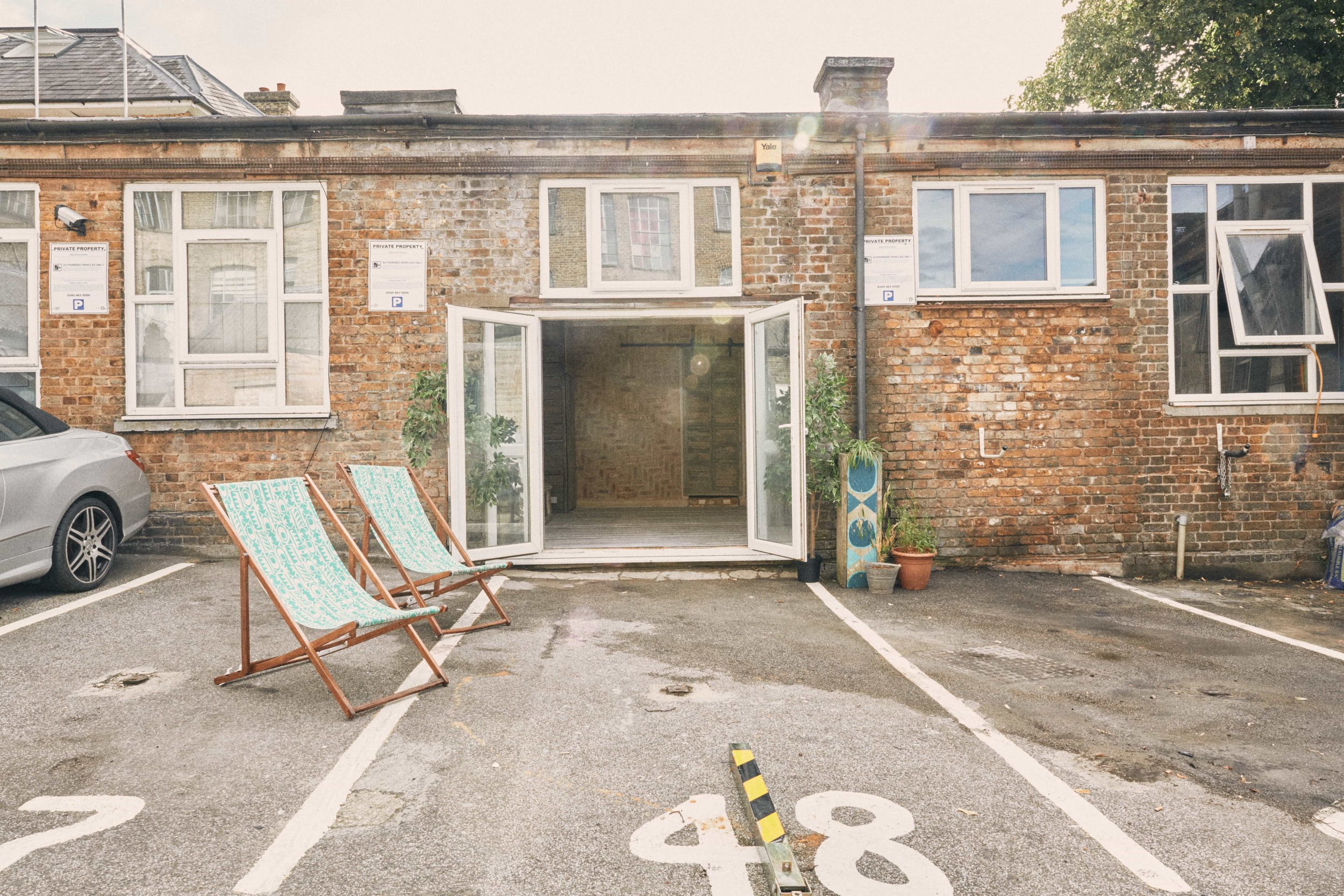 A brick apartment building with a glass door opening to a patio area with two deck chairs and several potted plants. The apartment has multiple windows, some open, and parking spaces numbered 48 and 49 in front.