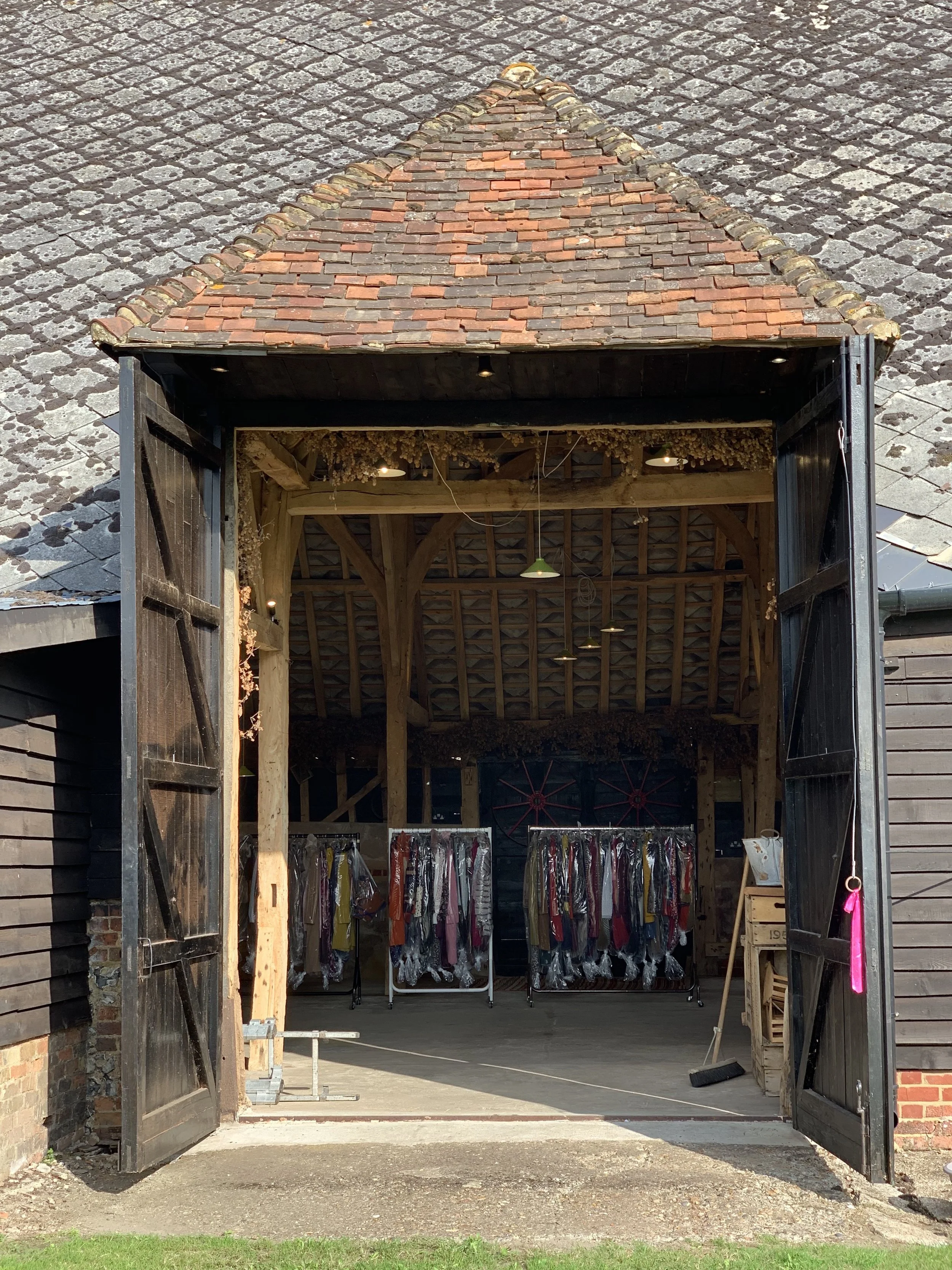 Open barn door revealing a clothing store with dresses hanging on racks inside, wooden interior, and a tiled roof visible from the outside.