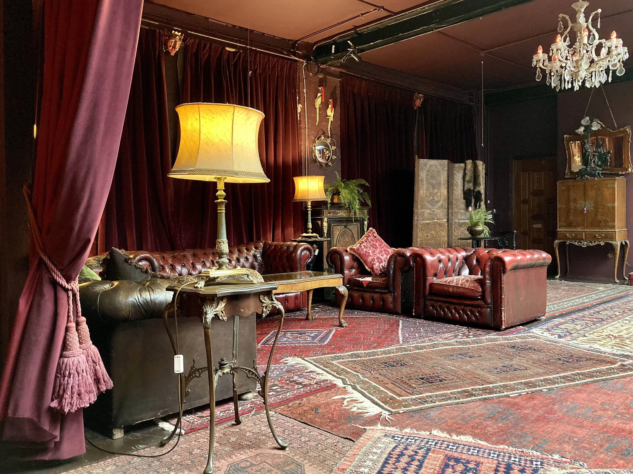 Vintage living room with red leather chesterfield sofas, ornate lamps, Persian rugs, dark red curtains, floral decorations, and antique furniture.