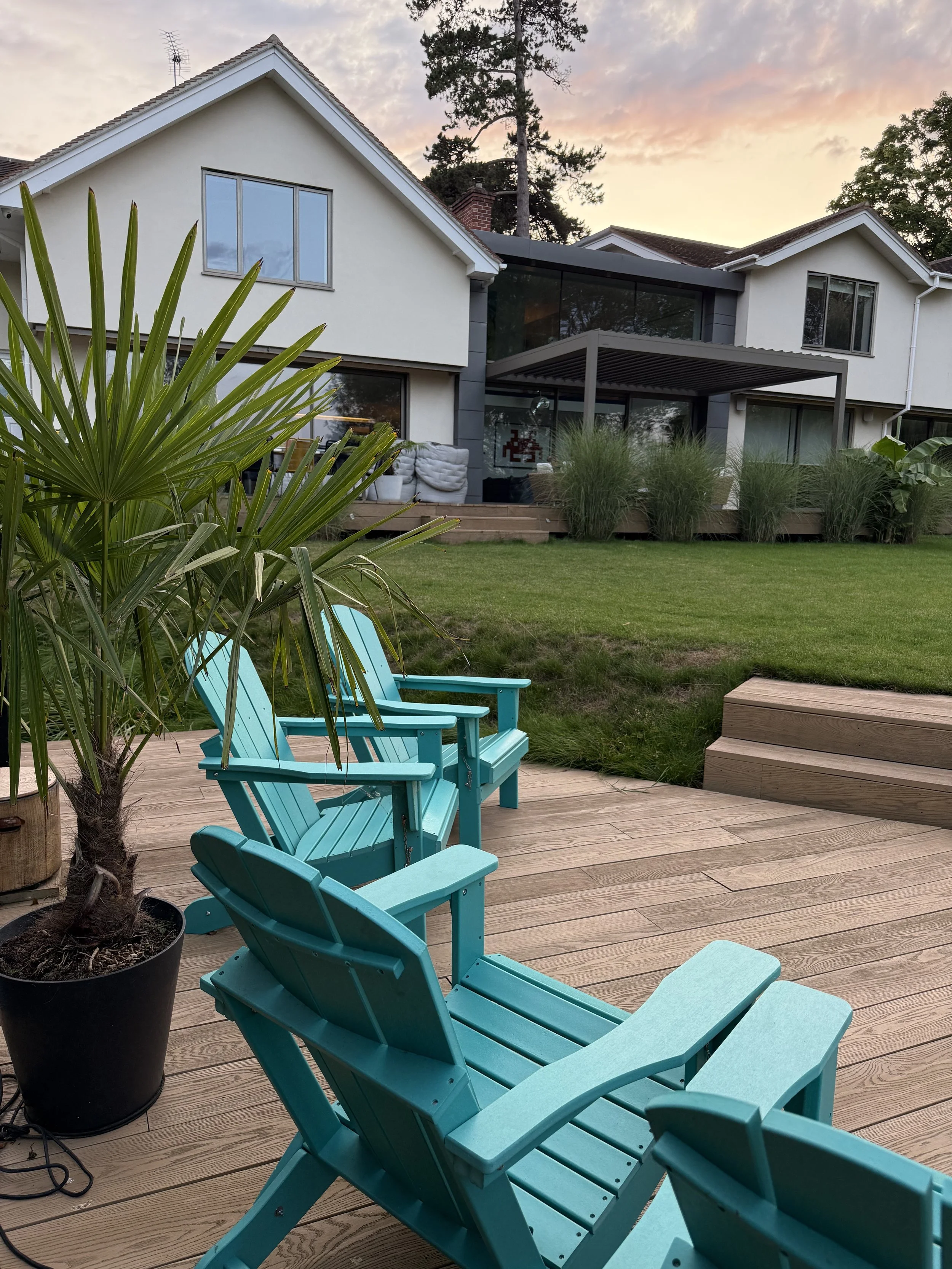 A backyard deck with three turquoise Adirondack chairs, a potted palm tree, and a grassy lawn leading to a modern house with large windows and a deck with seating, during sunset.