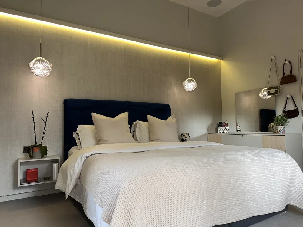 A modern bedroom with a large bed, white bedding, four pillows, a blue headboard, hanging globe pendant lights, a small side table with decorations, a large mirror, and wall-mounted bags and a plant.