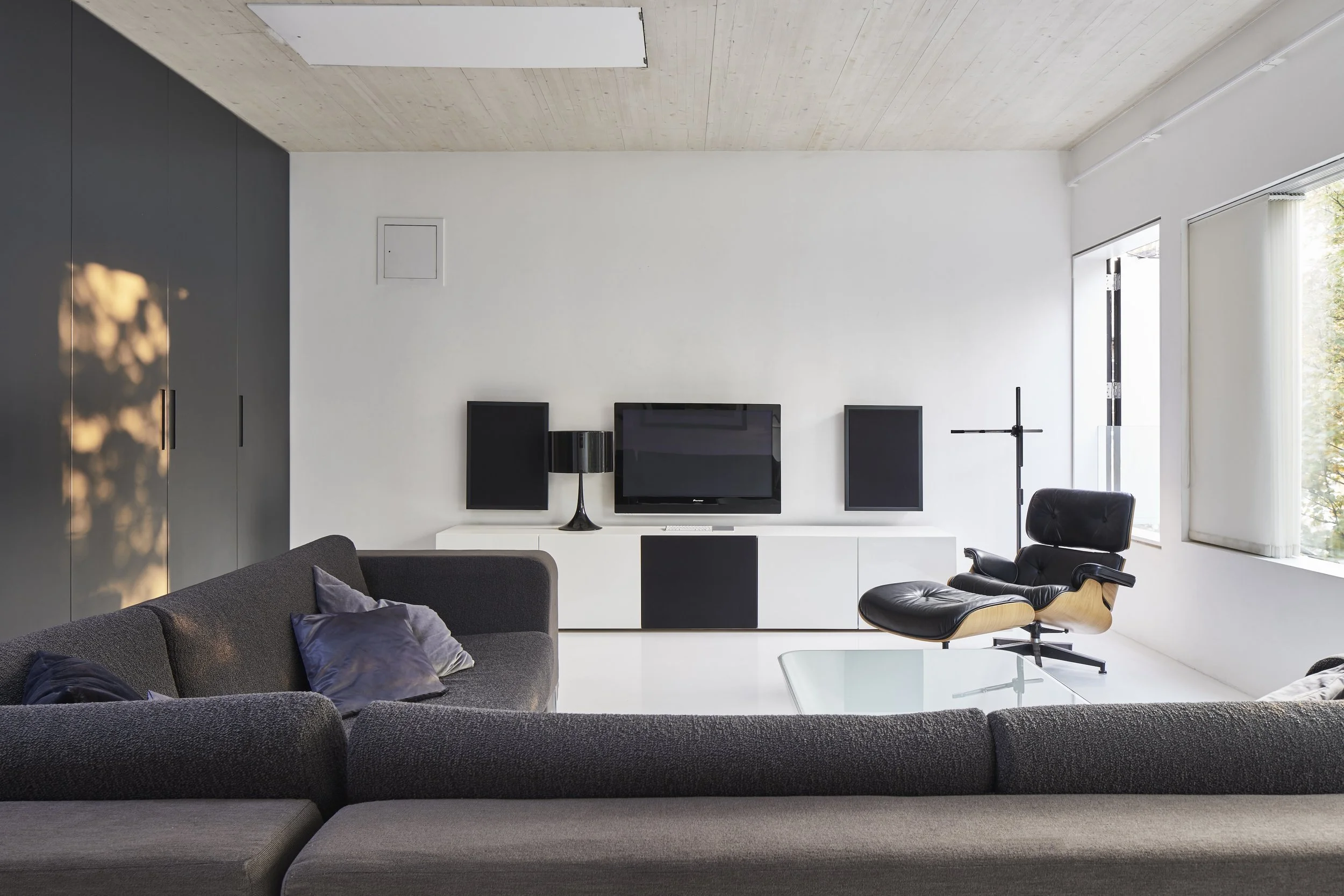 Modern living room with gray sofa, black lounge chair, TV mounted on white wall, black and white cabinet, window with blinds, and minimalist decor.