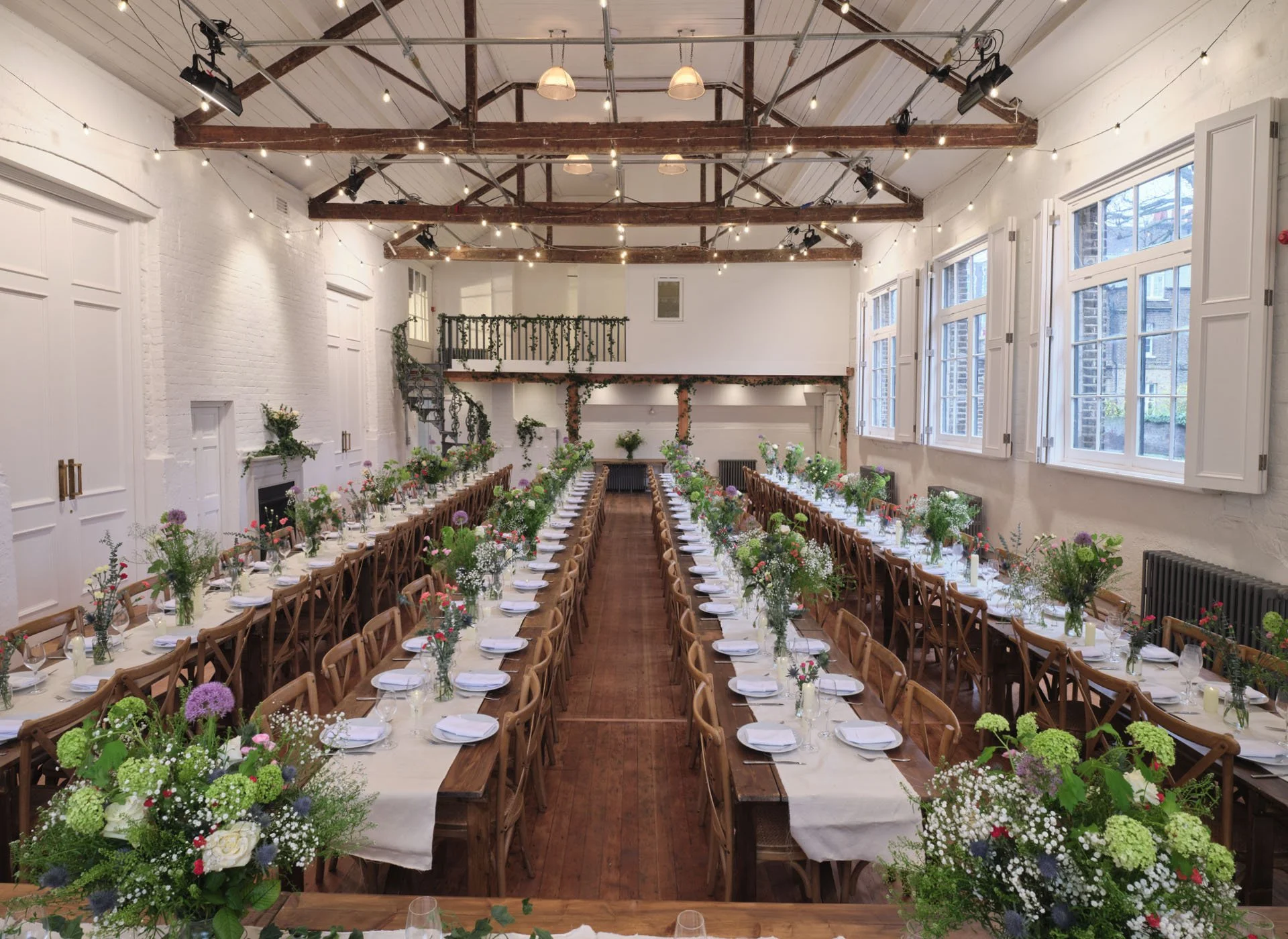 Long banquet-style tables set for an event in a bright, airy room with white brick walls, large windows, and wooden ceiling beams. The tables are decorated with white tablecloths, floral centerpieces, and place settings.