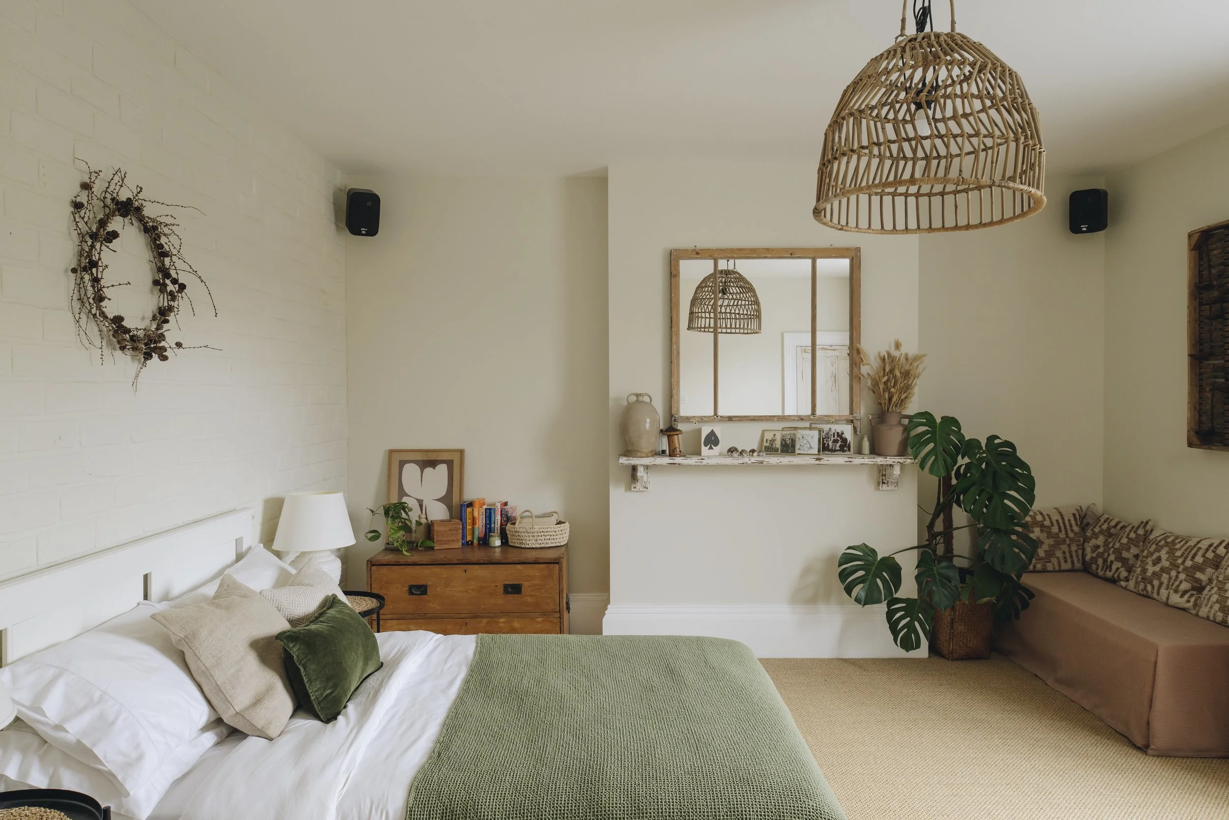 A cozy bedroom with a white brick wall, a bed with white and beige pillows, a green blanket, a wooden dresser with books and decorative items, a large mirror, a potted Monstera plant, a beige sofa with patterned cushions, and a wicker pendant light h