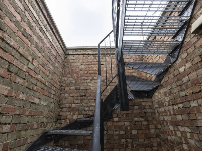 Black metal exterior staircase with three steps and guardrails, attached to a brick wall in an outdoor area.