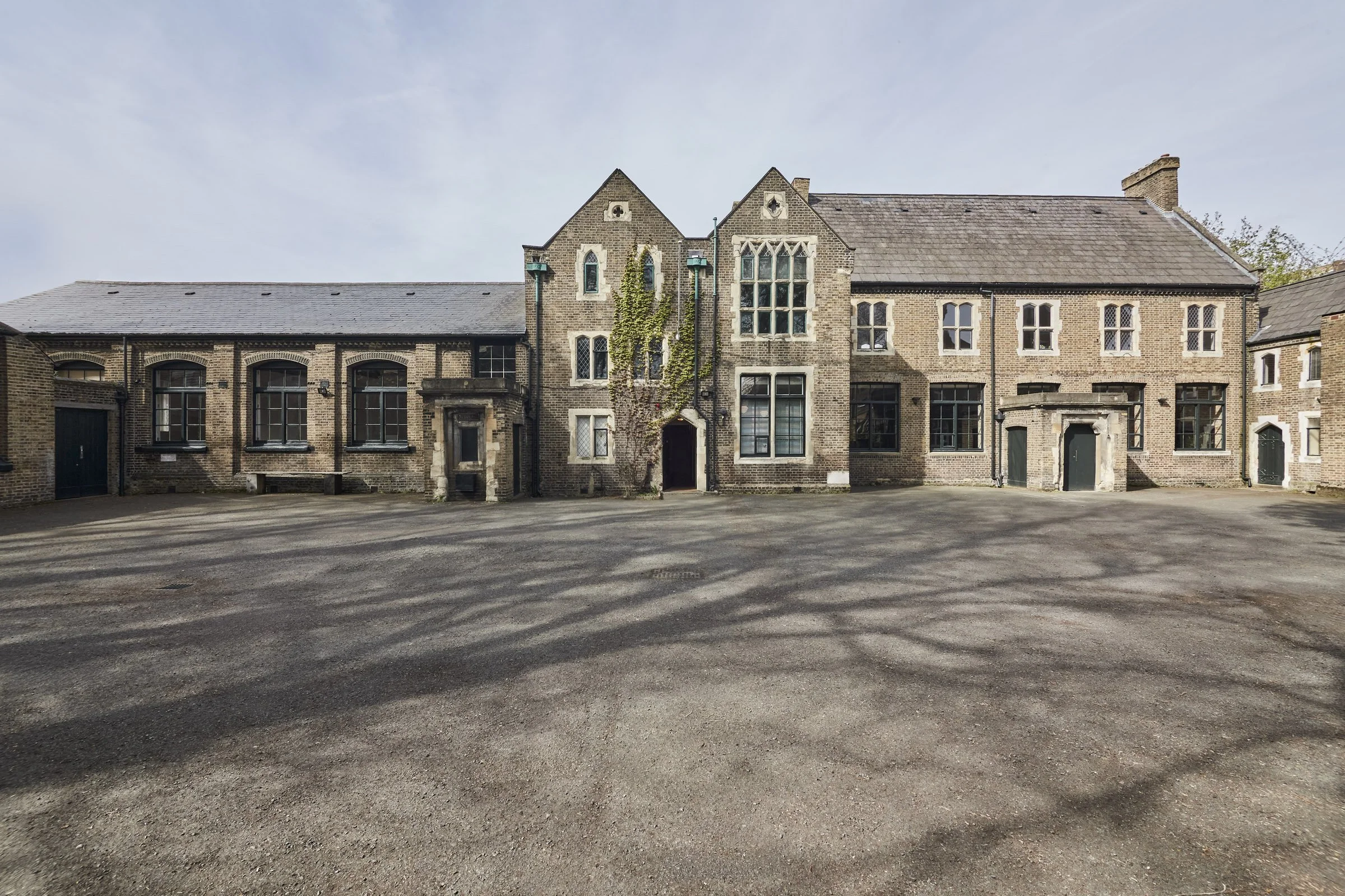 A large historic brick building with multiple windows and a central entrance, surrounded by an empty gravel courtyard.
