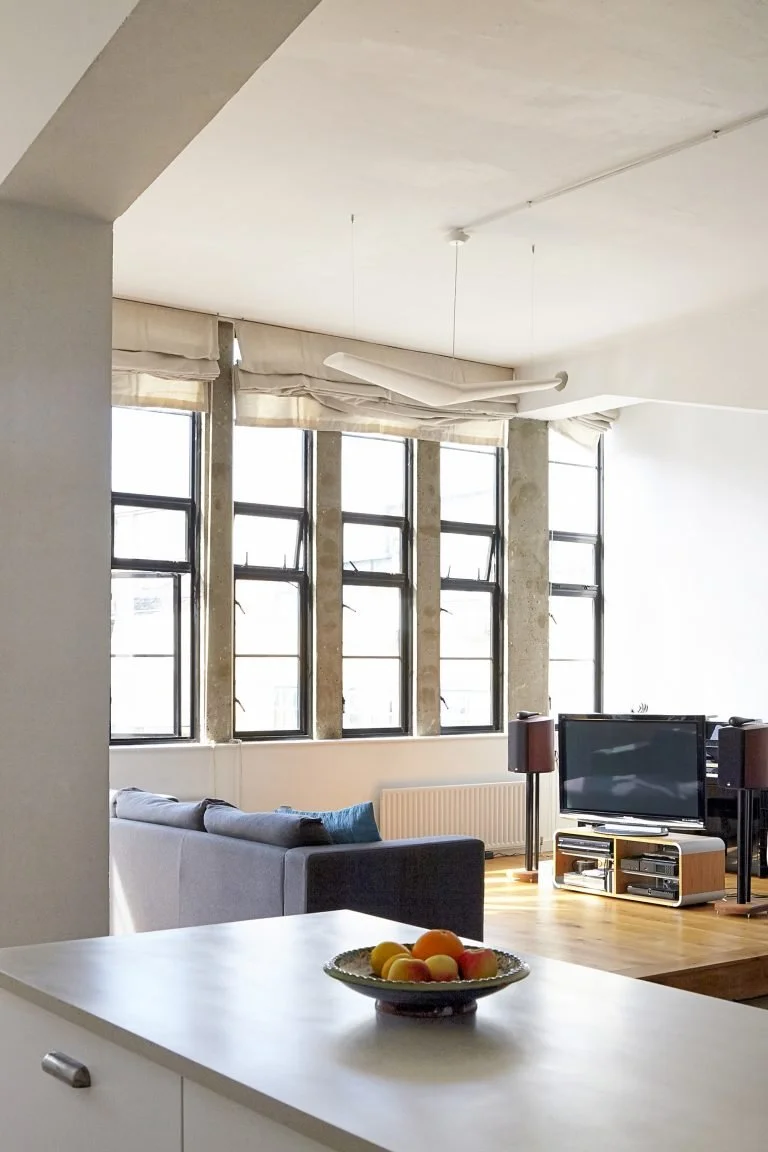 Bright living room with large windows, a gray sectional sofa, a television, and speakers, with a bowl of fruit on the white counter in the foreground.