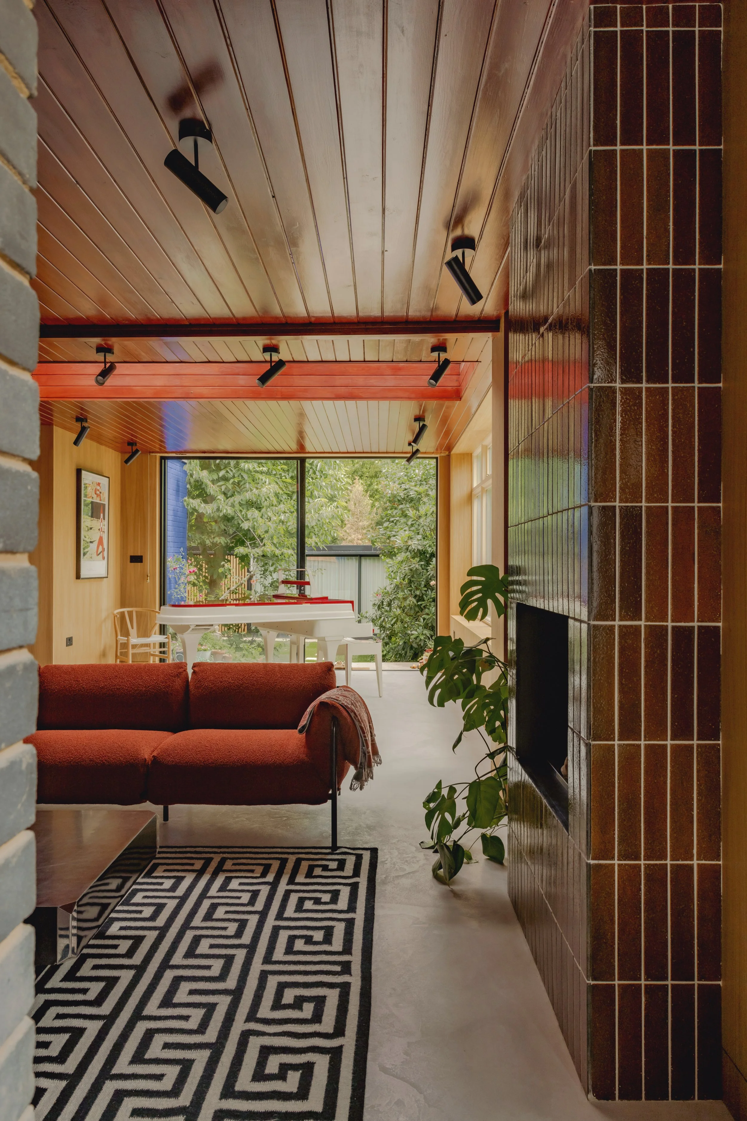 A living room with a maroon sofa, a black and white geometric rug, and a tall green plant next to a tiled wall. In the background, a white piano and chairs are visible near large windows showing greenery outside. The room has wooden ceiling panels wi