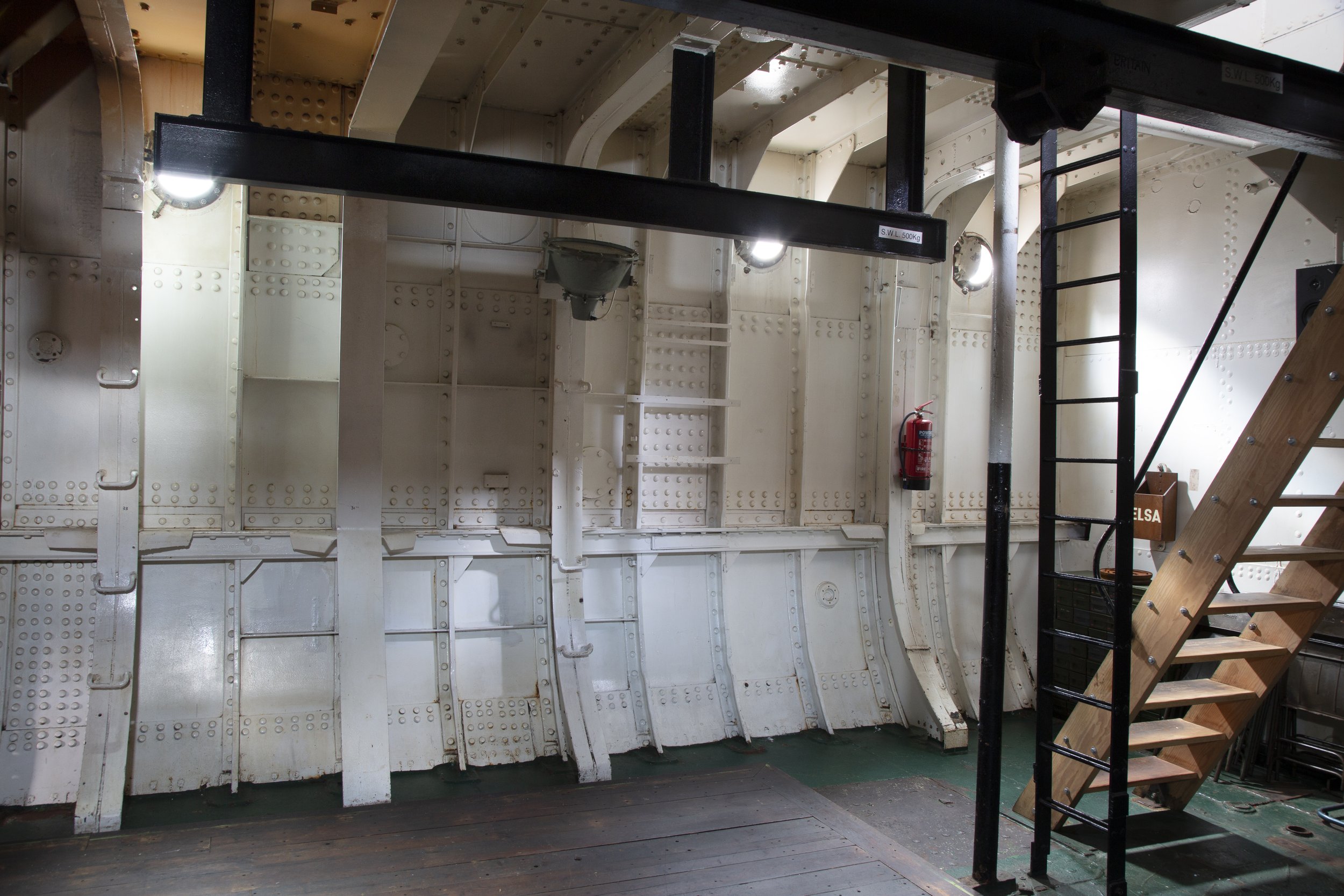 Interior of a ship or submarine with white painted metal walls, a fire extinguisher mounted on the wall, and a wooden staircase with metal railing leading upward.
