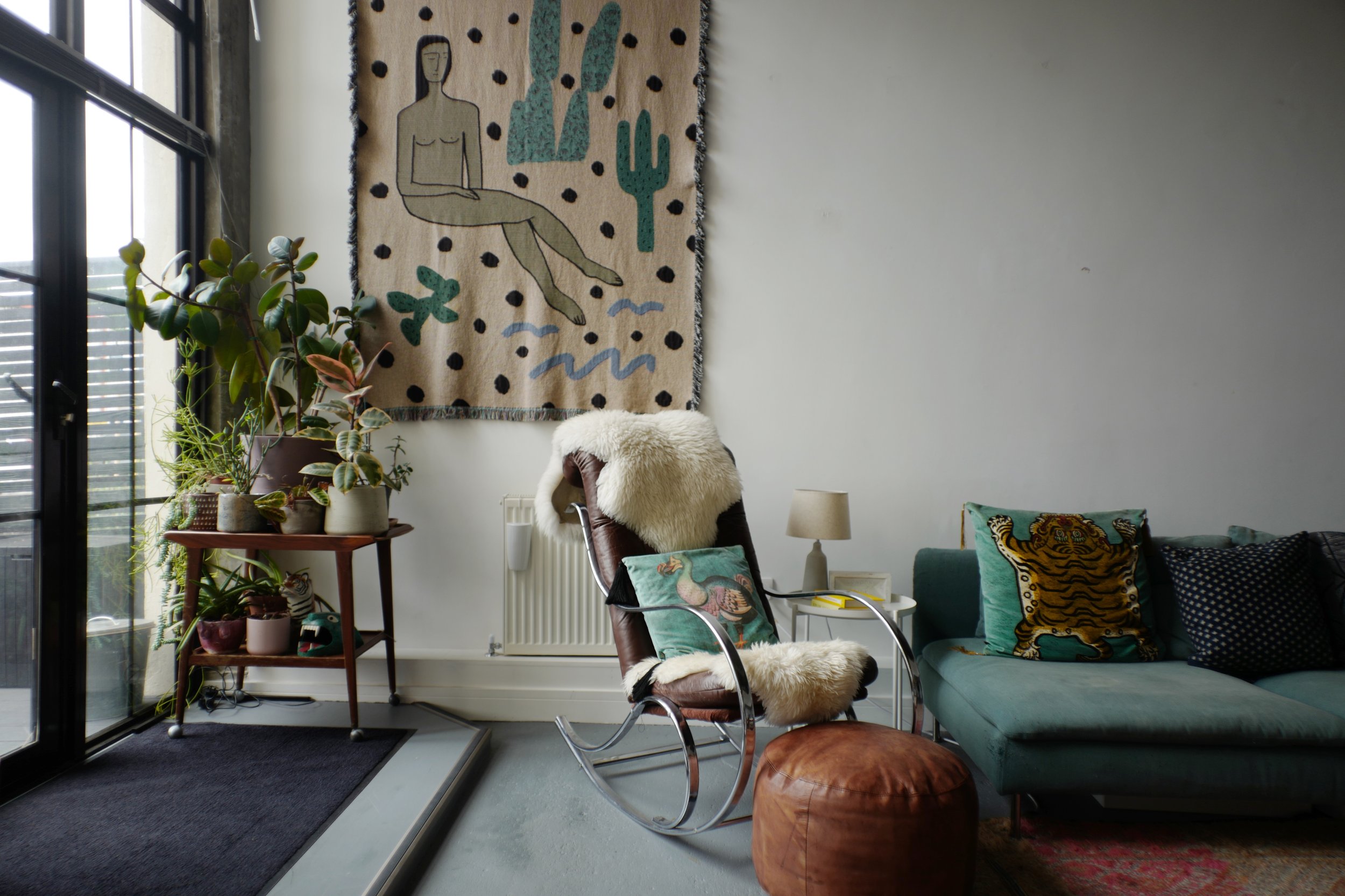 Living room with a glass door leading outside, potted plants on a small table, a wall hanging with an illustration of a woman and cacti, a brown rocking chair with a fluffy throw and a pillow, a small side table with a lamp, a teal couch with decorat