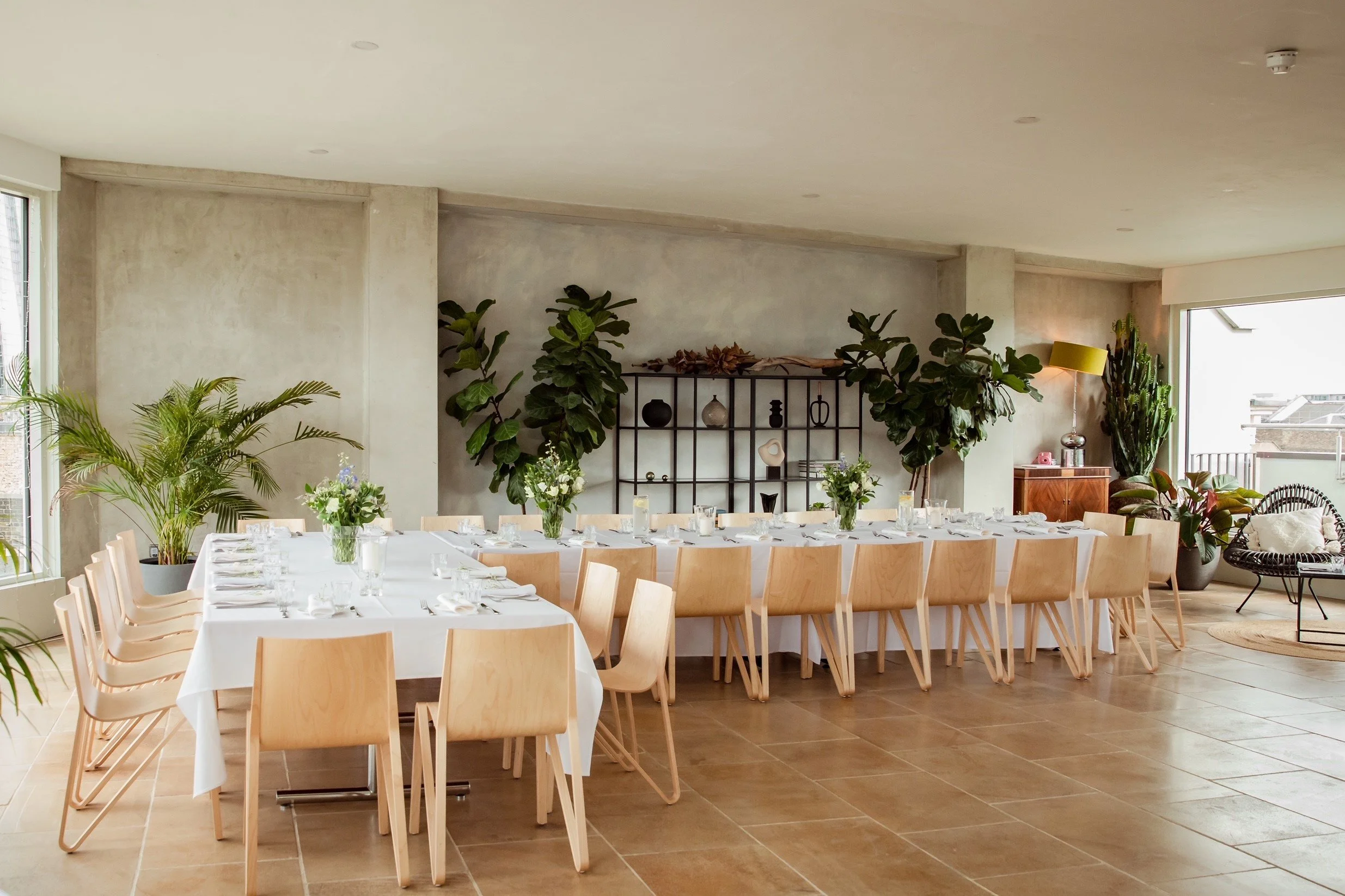 Modern dining room set up for a gathering with a long table covered with white tablecloths, set with glasses, plates, and utensils, surrounded by wooden chairs, decorated with floral arrangements, and featuring large potted plants and a shelving unit