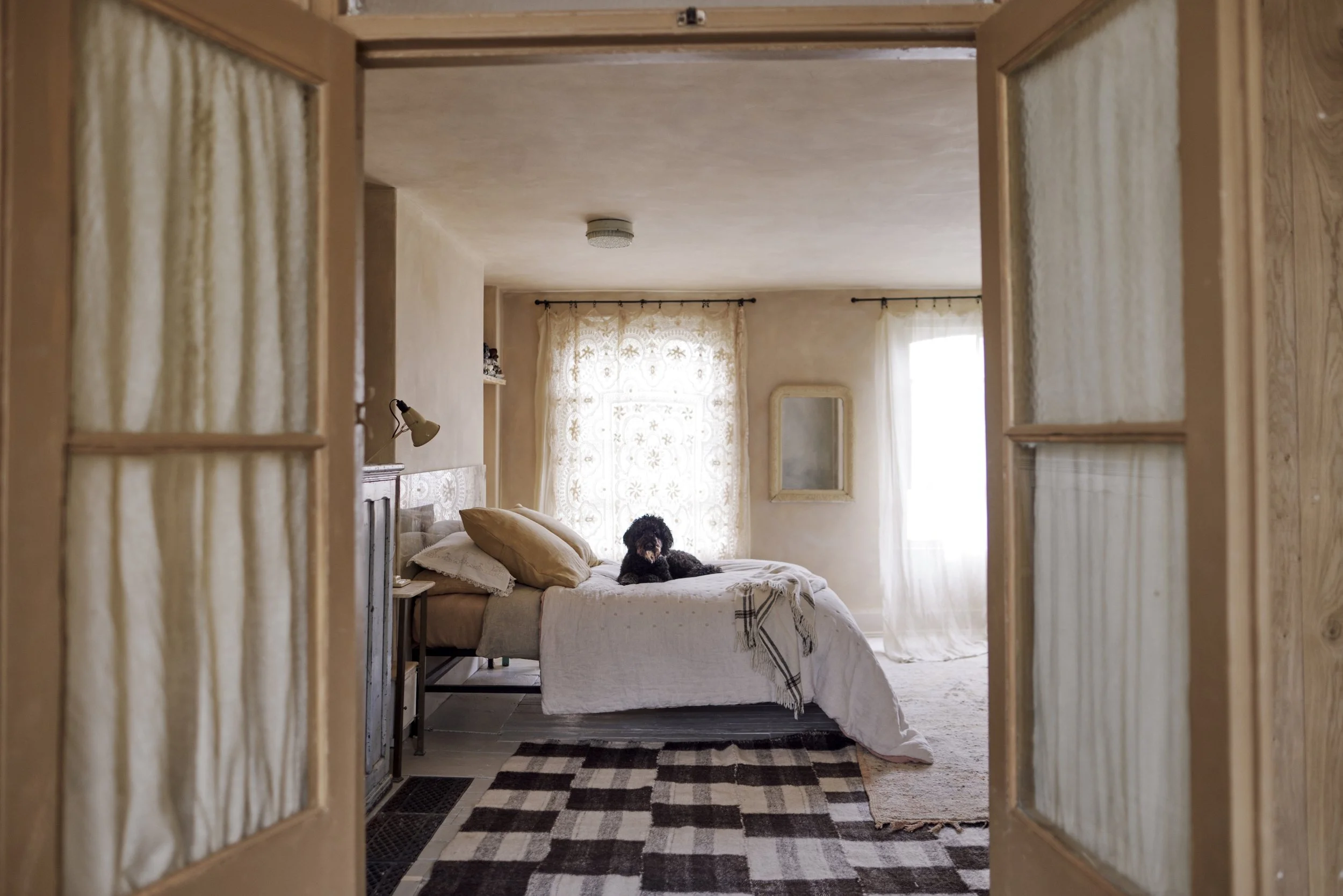 A cozy bedroom seen through a wooden door frame, featuring a bed with beige and white bedding and pillows, a black dog lying on the bed, large curtains with lace pattern, a mirror on the wall, and a checkered rug on the floor.