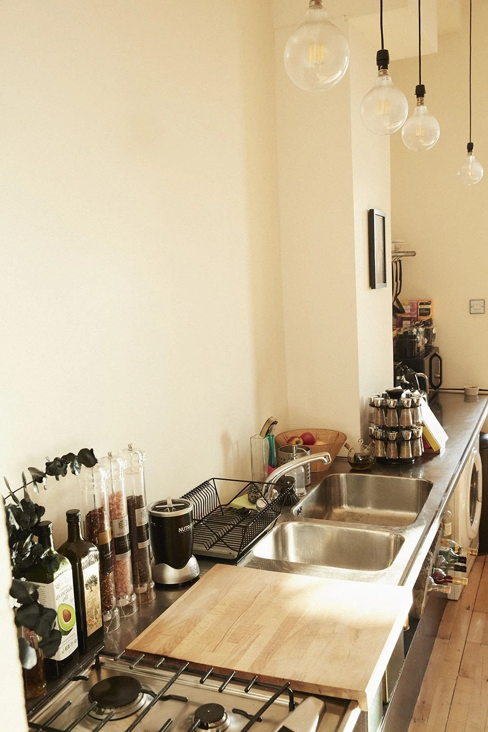 Kitchen countertop with two stainless steel sinks, a cutting board, spice jars, a coffee station with a grinder, and overhead hanging light bulbs.