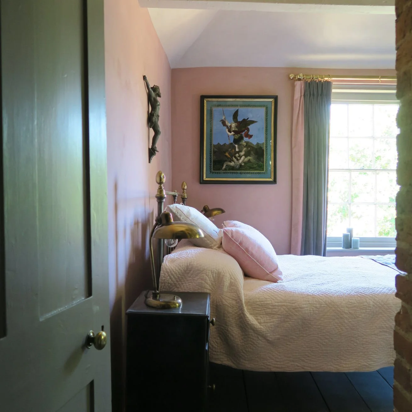 A cozy bedroom with pink walls, a large window with blue and pink curtains, a bed with a white quilt and pink pillows, a small black nightstand, and a bronze lamp. A framed painting and a metal wall sculpture are hanging on the wall.