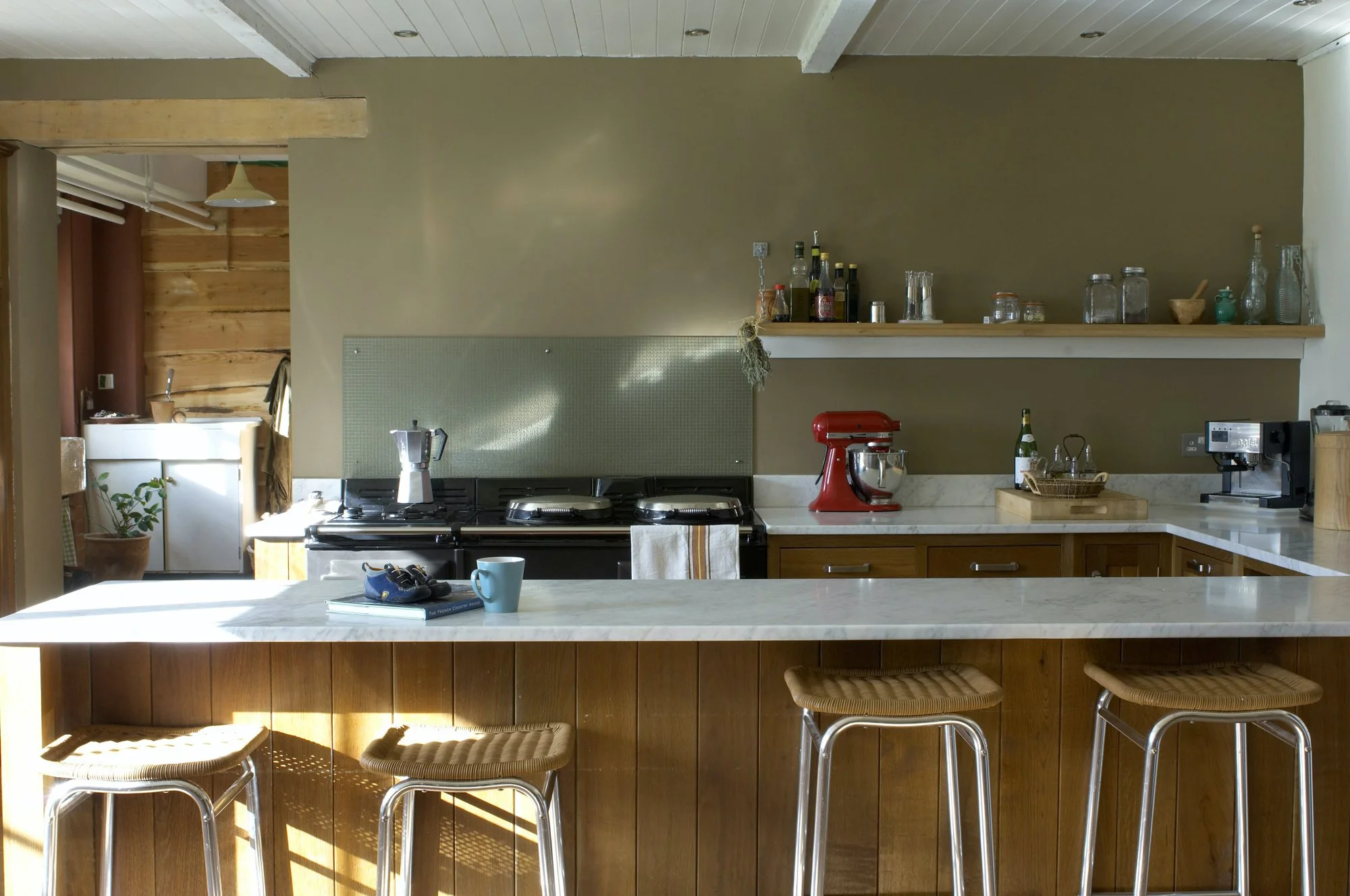 Kitchen with a white marble island, three stools, and a view of the stove, cabinets, and open shelves with bottles, jars, and small containers.