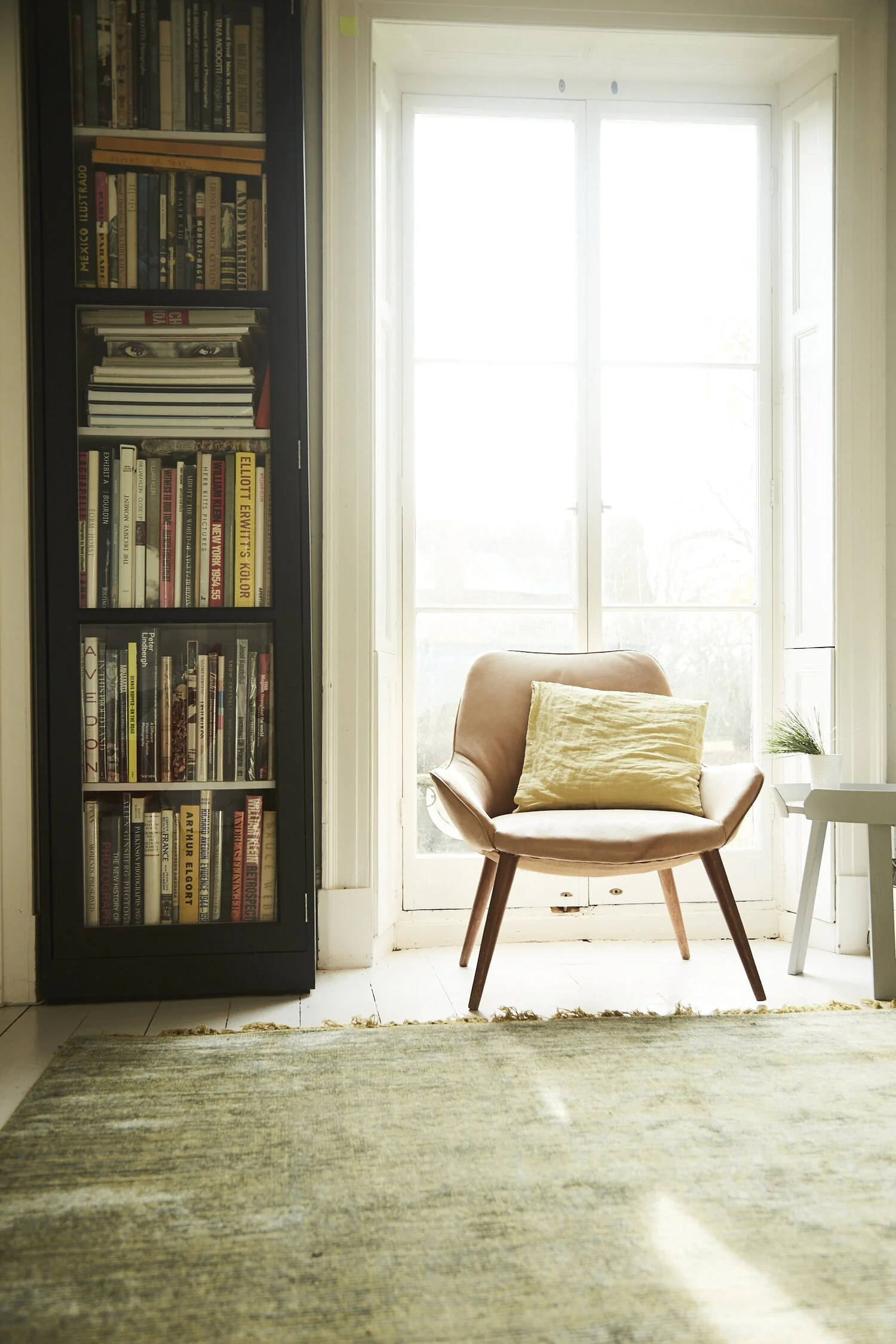 A cozy corner with a pink armchair with a yellow pillow, next to a small white table with a potted plant, in front of a large window with bright daylight, and a black bookshelf filled with books on the left, and a green rug on the floor.