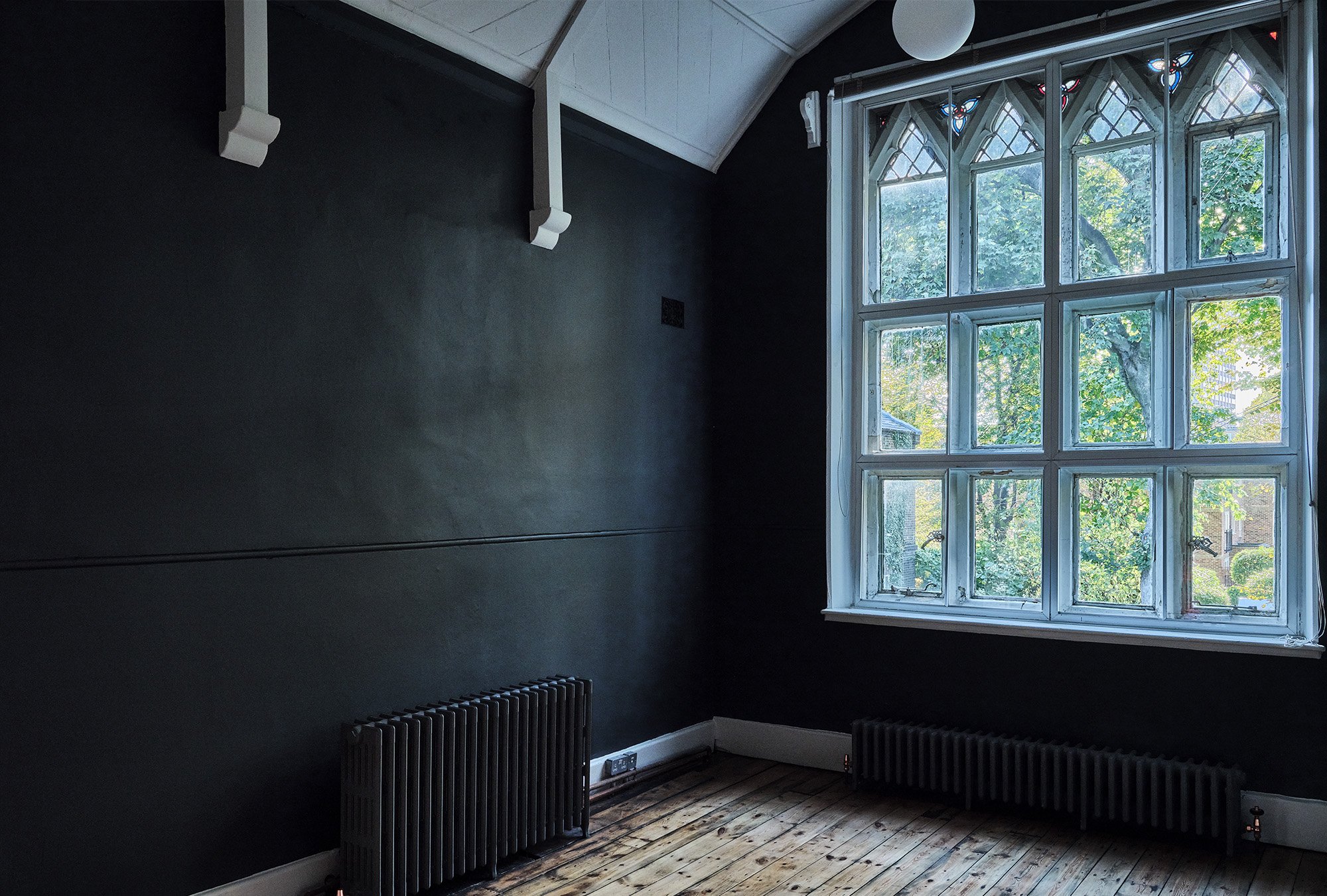 Interior of a room with dark walls, a large Gothic-style window, wooden floor, and radiators beneath the window.