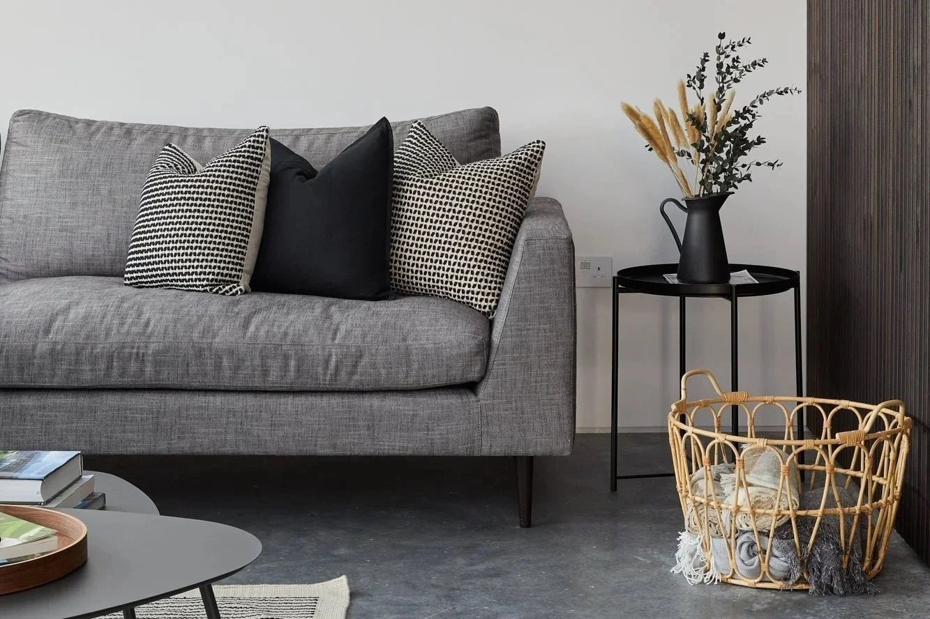 Living room with a grey sofa, decorative pillows, a black side table with a black vase and dried plants, a wicker basket with rolled towels, and a black textured wall panel.