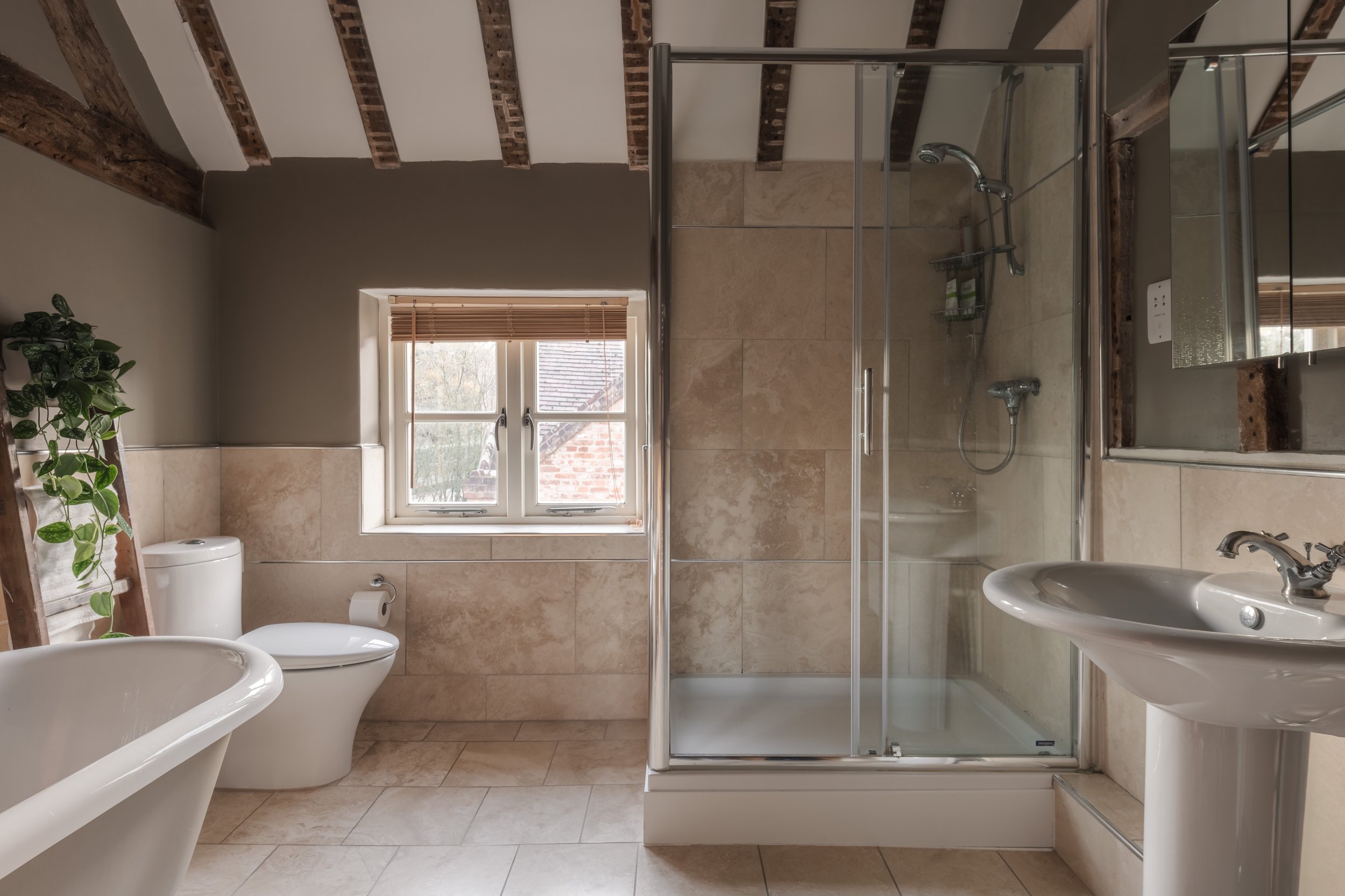A bathroom with a shower, toilet, pedestal sink, clawfoot bathtub, window with blinds, and exposed brick ceiling beams.