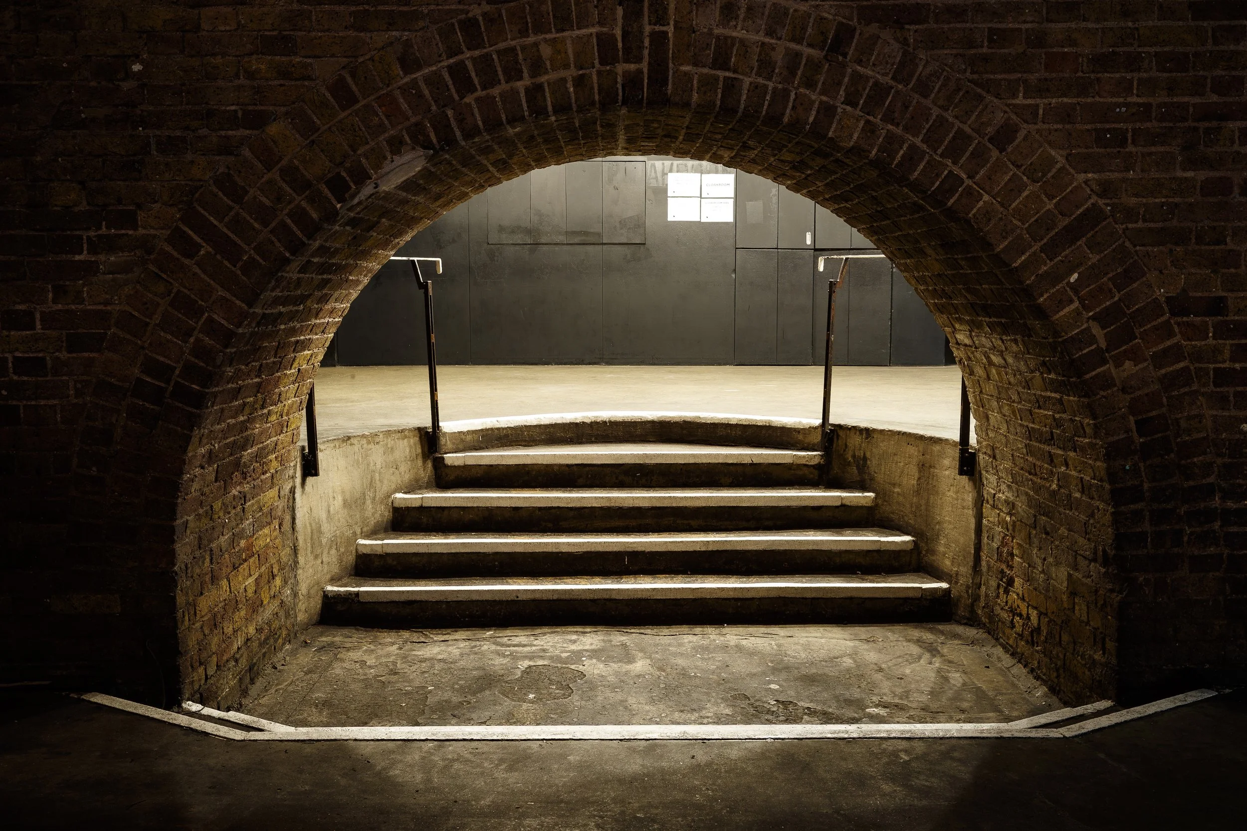 Brick tunnel opening leading to stairs with metal handrails and a black wall at the top.