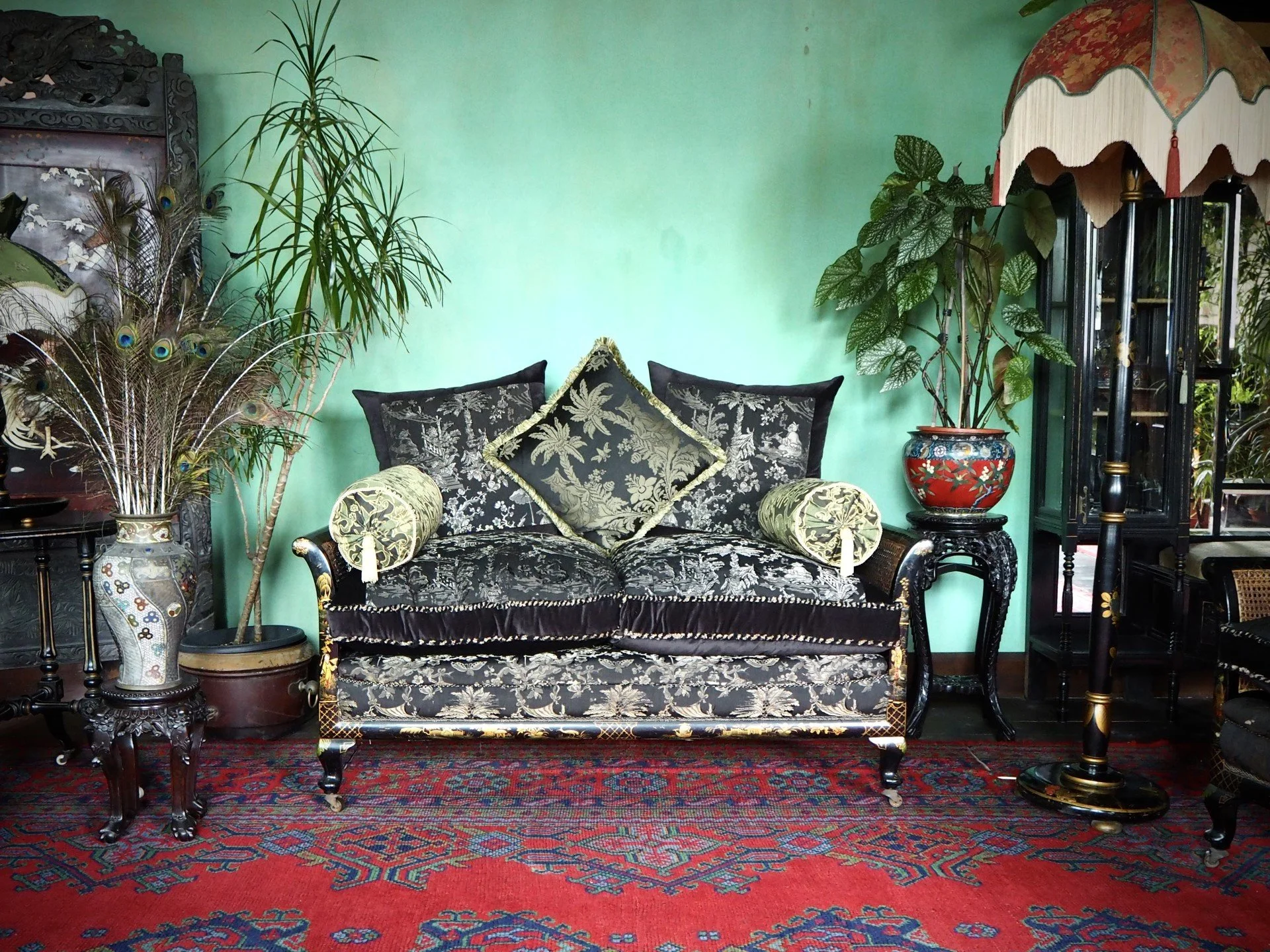 An elaborately decorated antique-style living room with a black velvet sofa with gold floral patterns, green cushions, and matching bolsters. Potted plants and peacock feathers in a vase are on the left, and a tall glass display cabinet with decorati