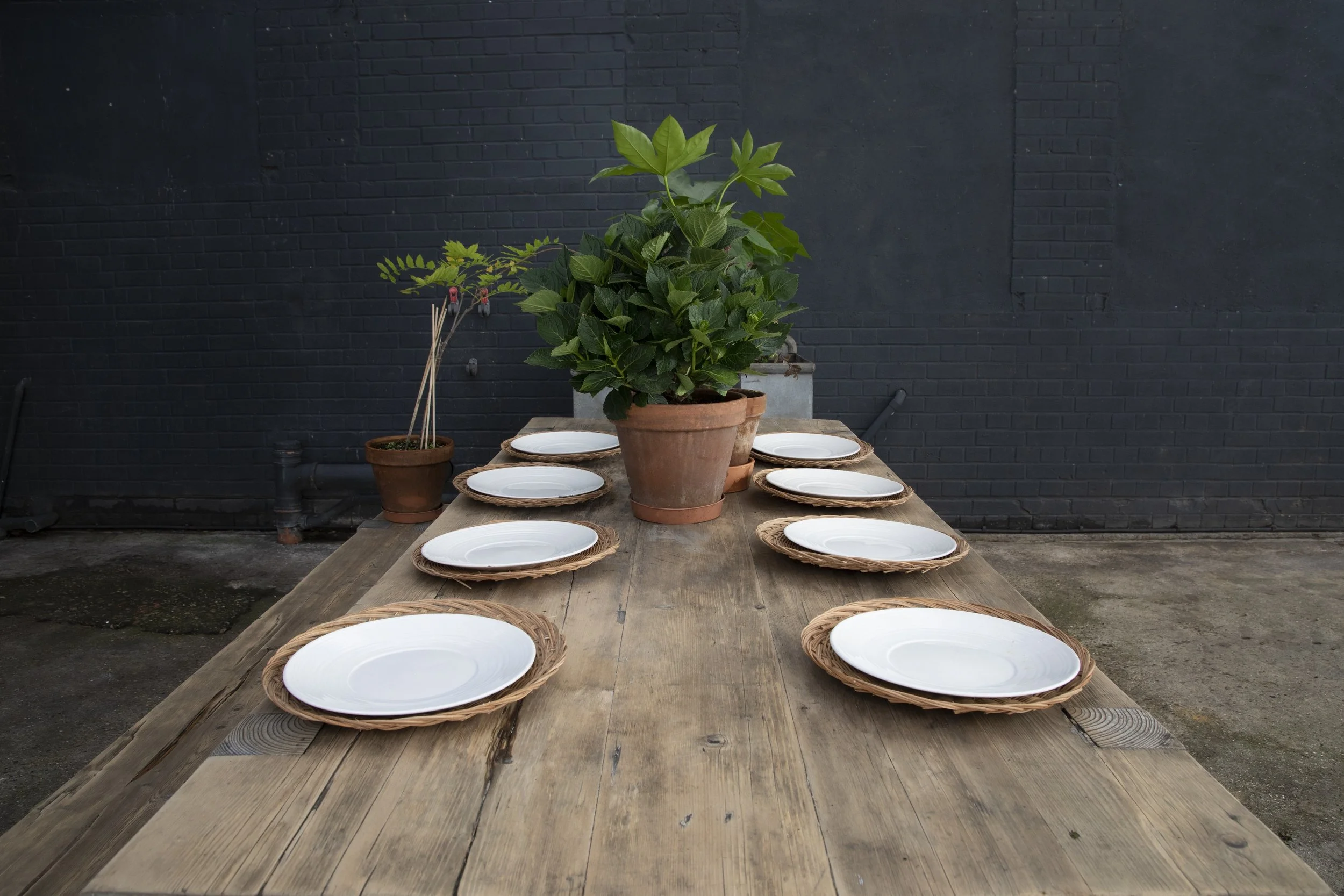 An outdoor wooden dining table set with eight white plates on wicker placemats, brown potted plants, and a black brick wall in the background.