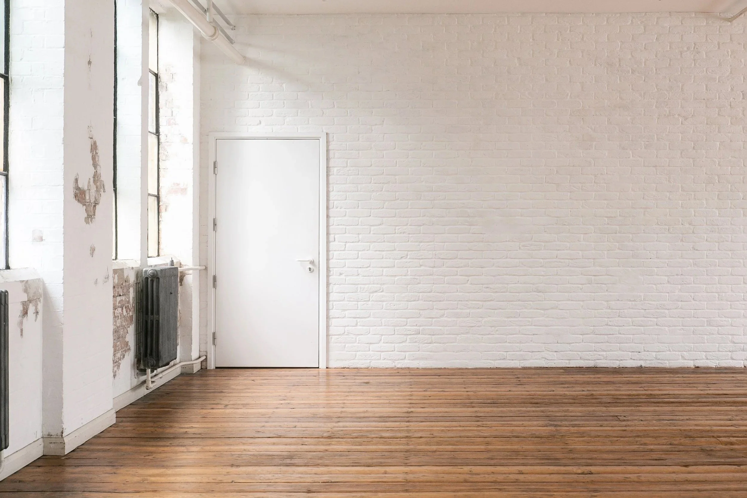 Empty room with white brick walls, wooden floor, and large windows, some with visible peeling paint.